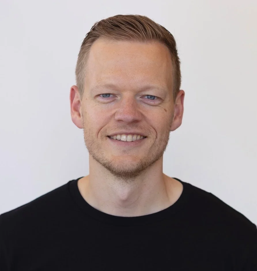 A headshot of a smiling man with short, light brown hair, blue eyes, and light facial hair, wearing a black shirt against a plain white background.