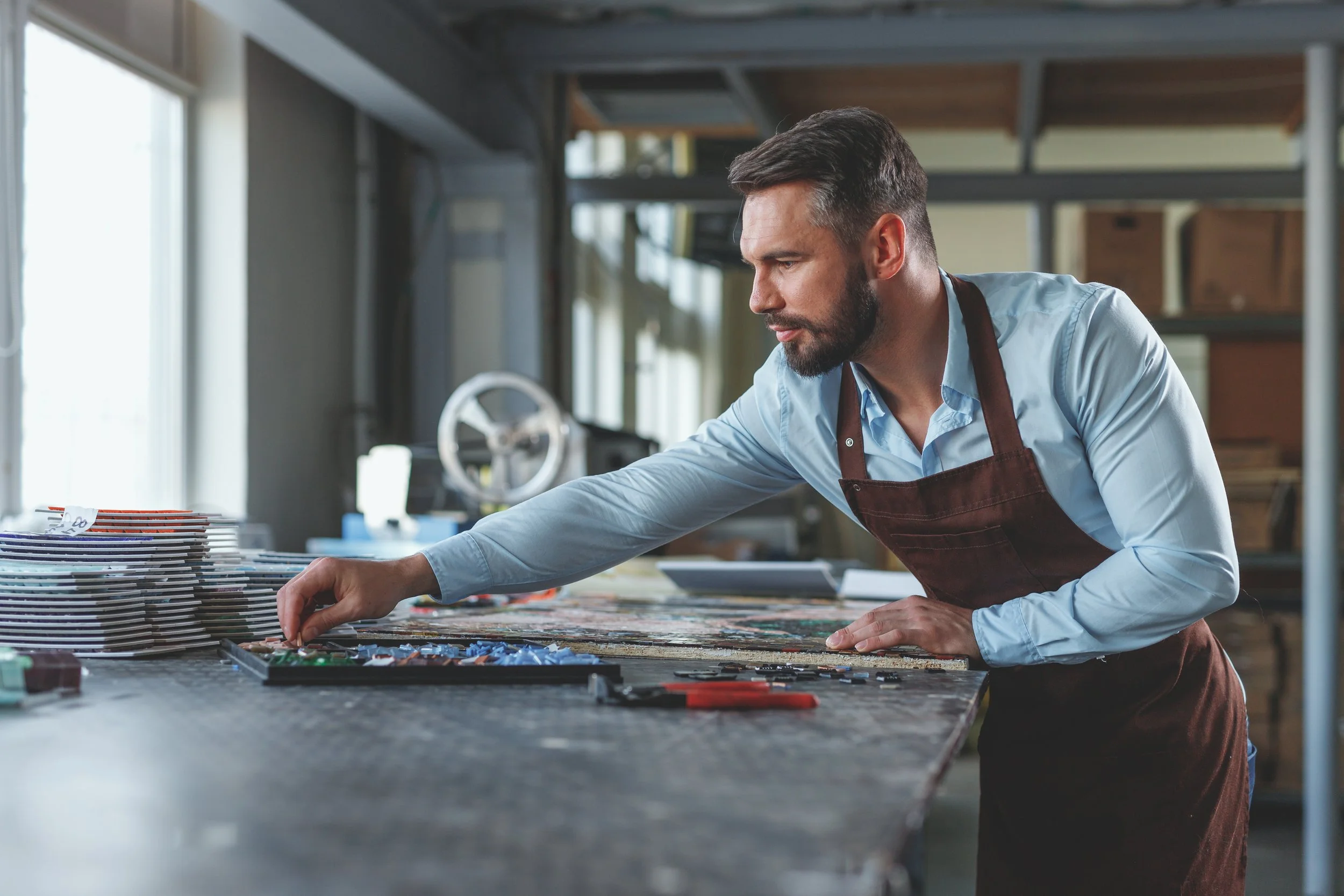 Man working on a mosaic art project at a workshop table, arranging small tiles on a surface with stacks of papers or tiles nearby.