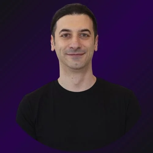 A man with dark hair, wearing a black shirt, smiling against a dark purple background.