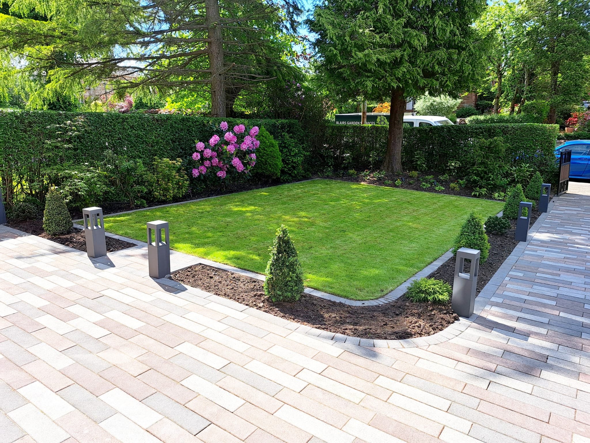 A neatly manicured small lawn with surrounding young shrubs and flowers, bordered by a paved walkway and modern outdoor lights, with lush green trees and shrubbery in the background.