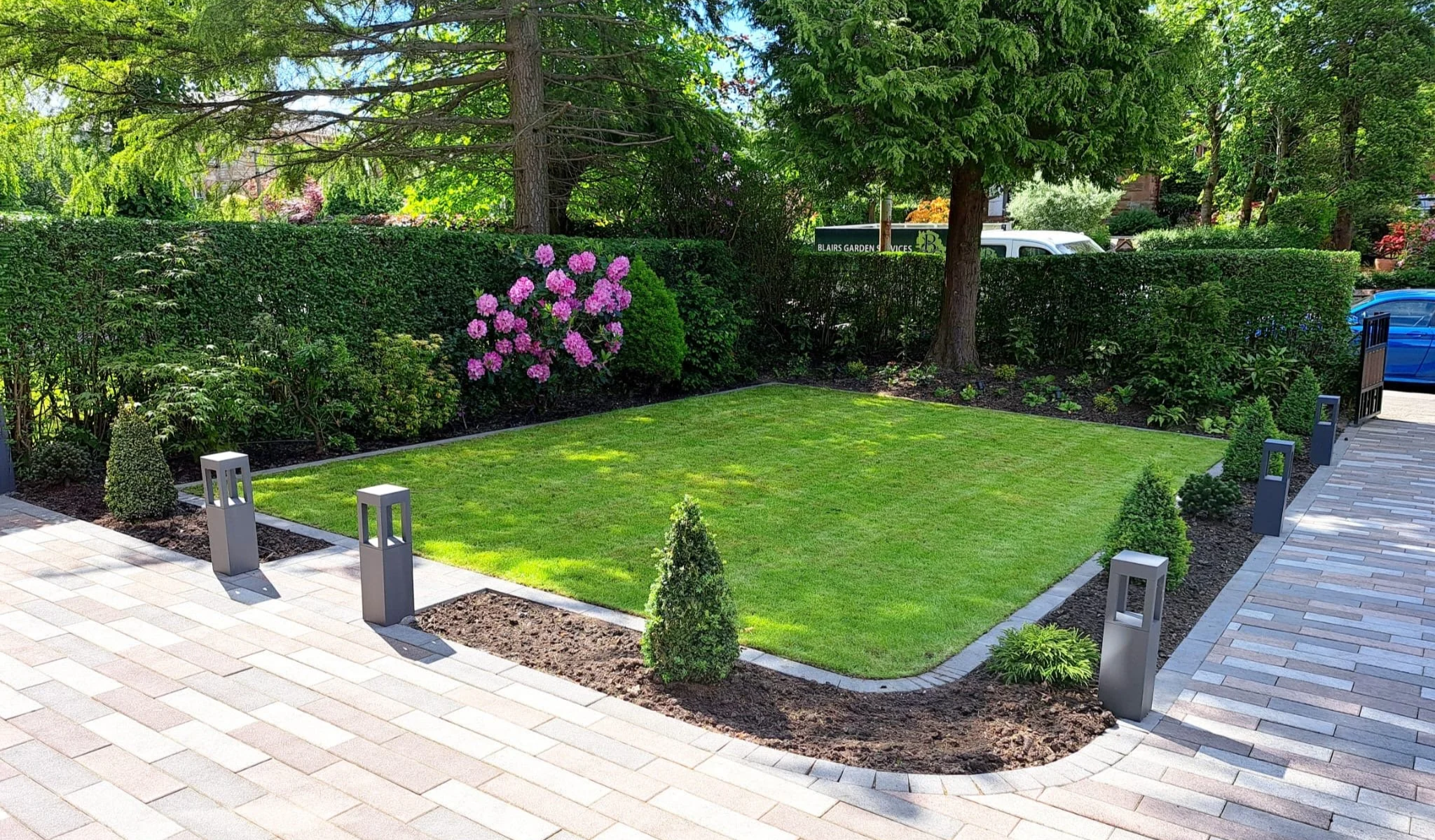 A small landscaped yard with a green grassy patch bordered by a paved walkway, small bushes, and flowers, with trees, shrubs, and a hedge in the background.