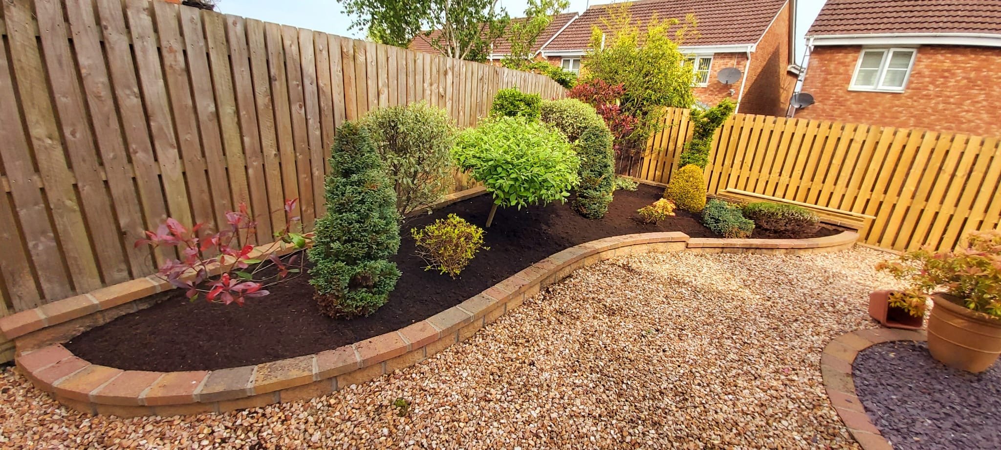Backyard garden with a variety of trimmed bushes and small trees, bordered by a curved brick wall and a wooden fence, with a gravel pathway and a potted plant.