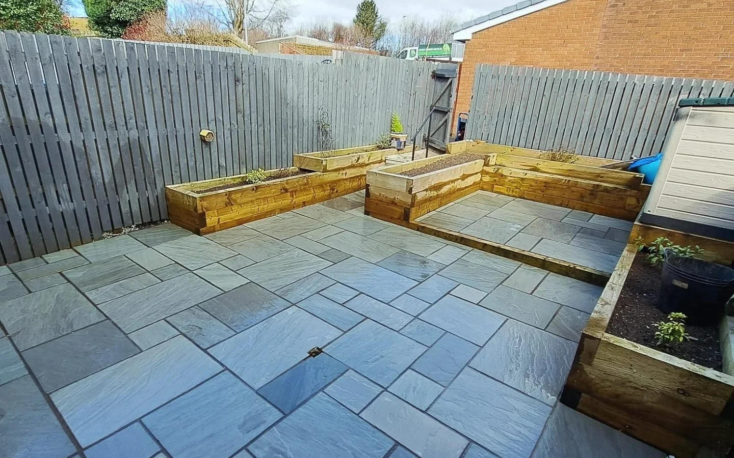 A backyard patio with blue-gray stone tiles, surrounded by wooden raised garden beds and a gray wooden fence.