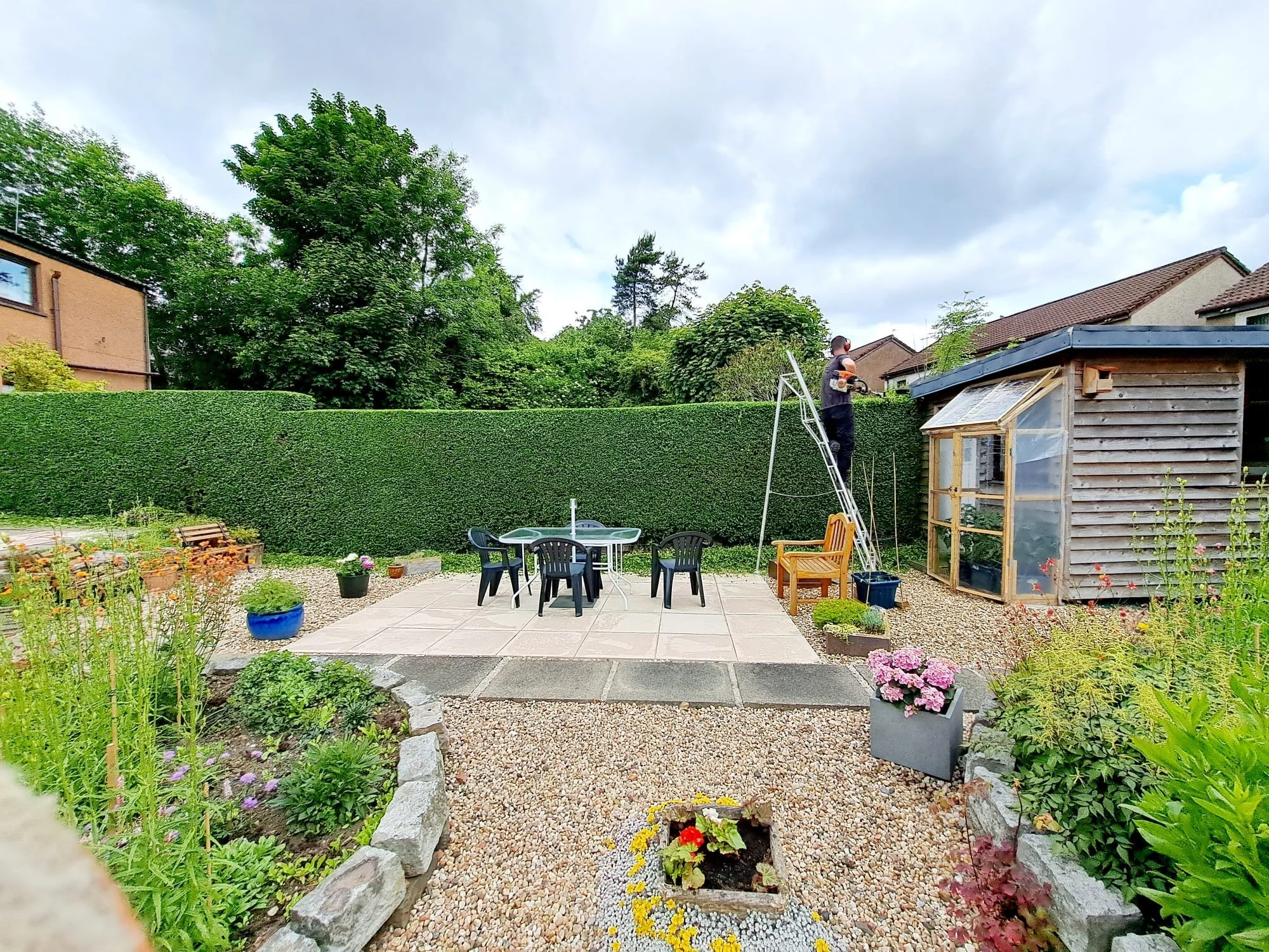 A backyard with a landscaped garden, a round table with black chairs on a tiled patio, a shed, a person on a ladder trimming a tall hedge, and various potted plants and flowers.