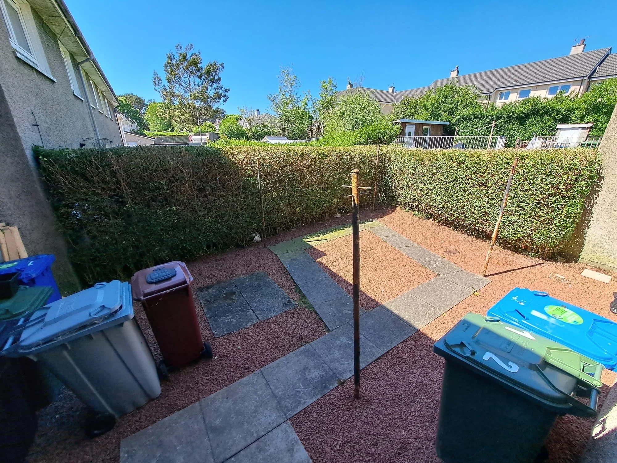 A small outdoor backyard with a paved stone path, surrounded by tall green hedges, under a clear blue sky. There are several trash and recycling bins on the side, and a few metal poles or clotheslines.