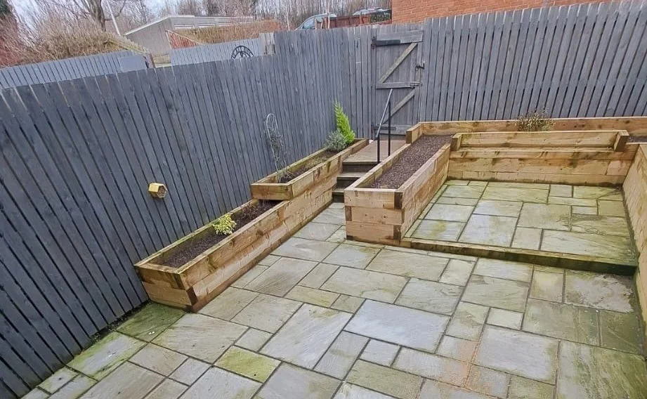 Small backyard patio with stone paving, raised wooden garden beds along a blue fence, small plants inside the beds, and a wooden trellis at the end.