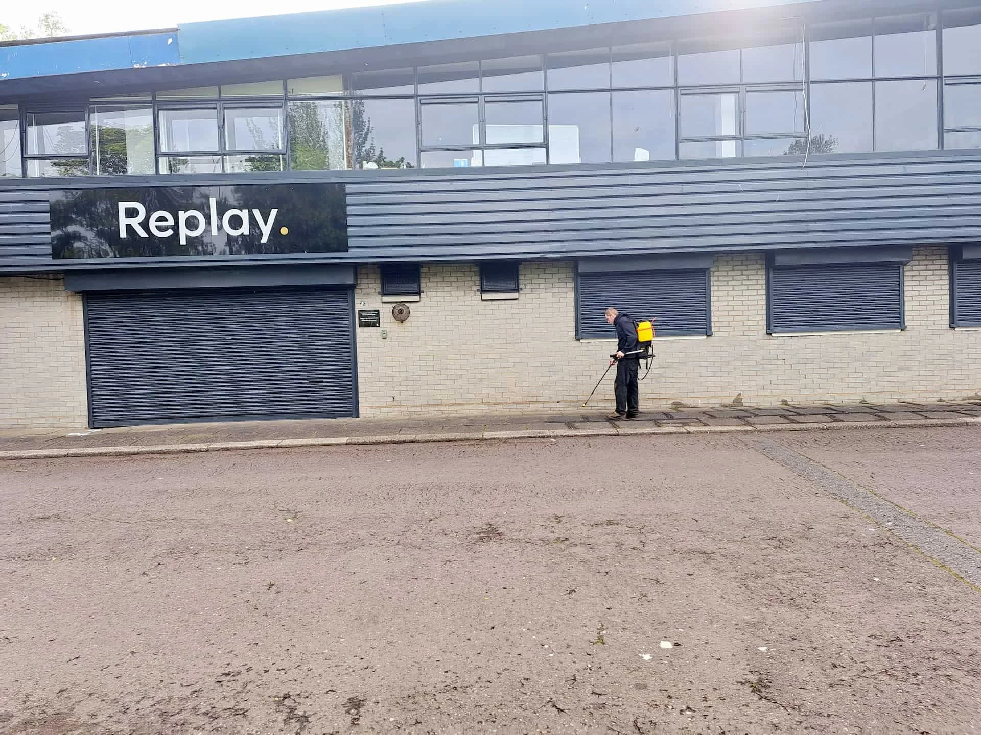 A person in dark clothing with a yellow backpack-style sprayer is pressure washing the sidewalk in front of a building with a black sign that says "Replay."