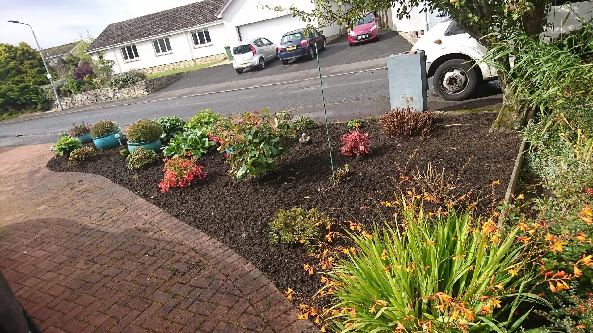 A landscaped front garden with a curved brick pathway, colorful plants, and potted plants along the edge, situated in a suburban neighborhood with houses, parked cars, and a tree.