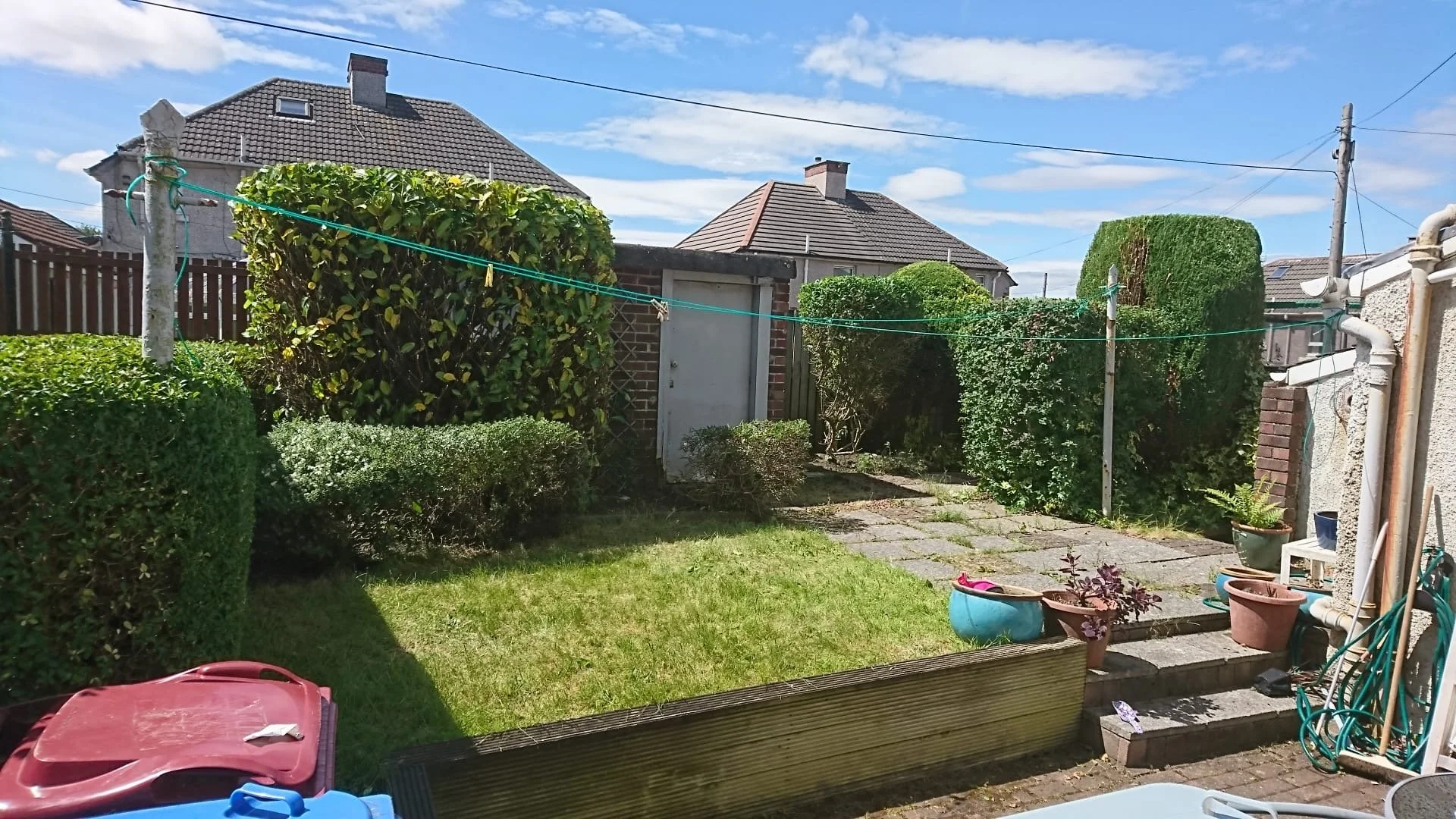 A backyard garden with trimmed bushes, a small patch of grass, potted plants, and a stone pathway. There are houses in the background under a blue sky with some clouds.