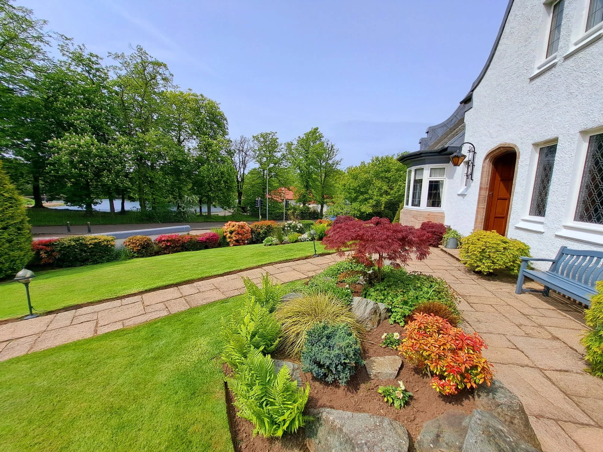A garden with grass, bushes, and a paved pathway alongside a white house with a brown door and multiple windows, surrounded by vibrant plants and trees under a blue sky.