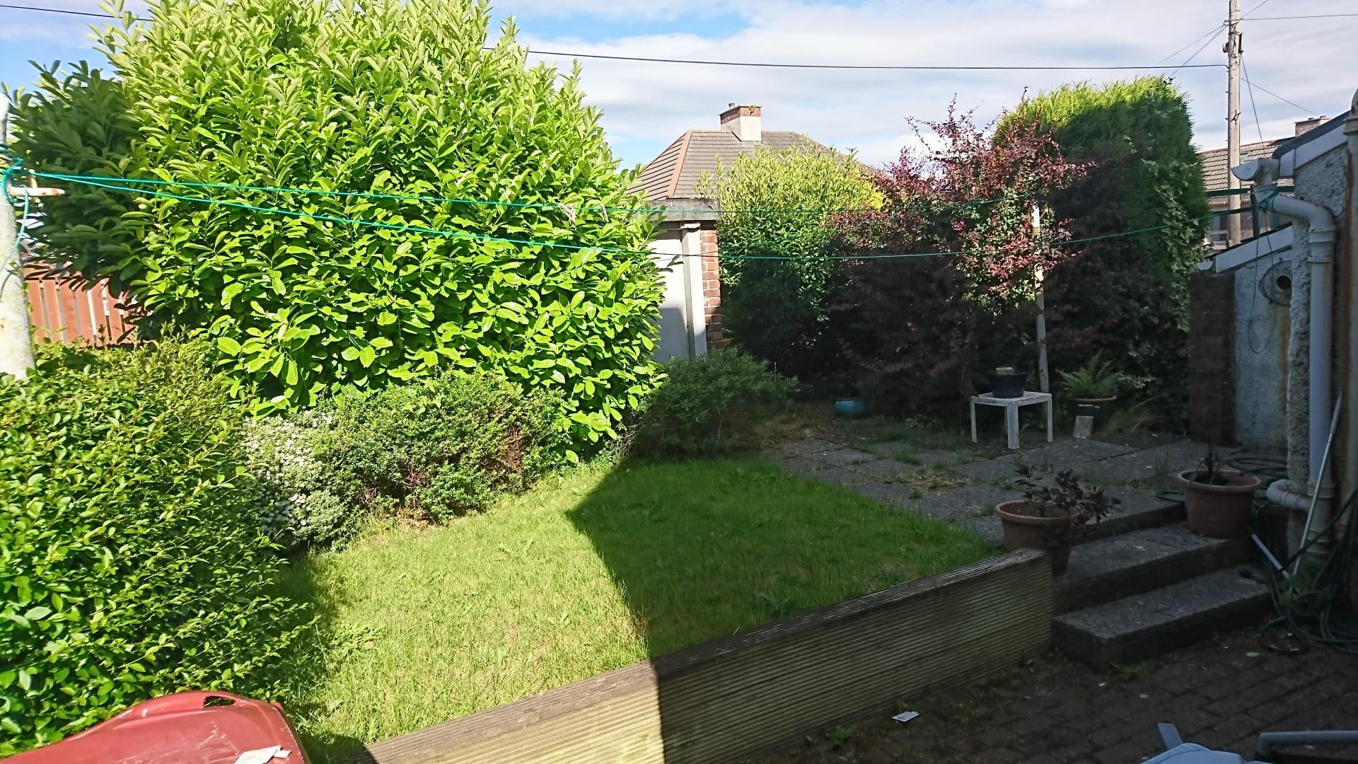 A backyard garden with a variety of lush green bushes and trees, a grassy area, and a paved patio with potted plants and outdoor furniture under a partly cloudy sky.
