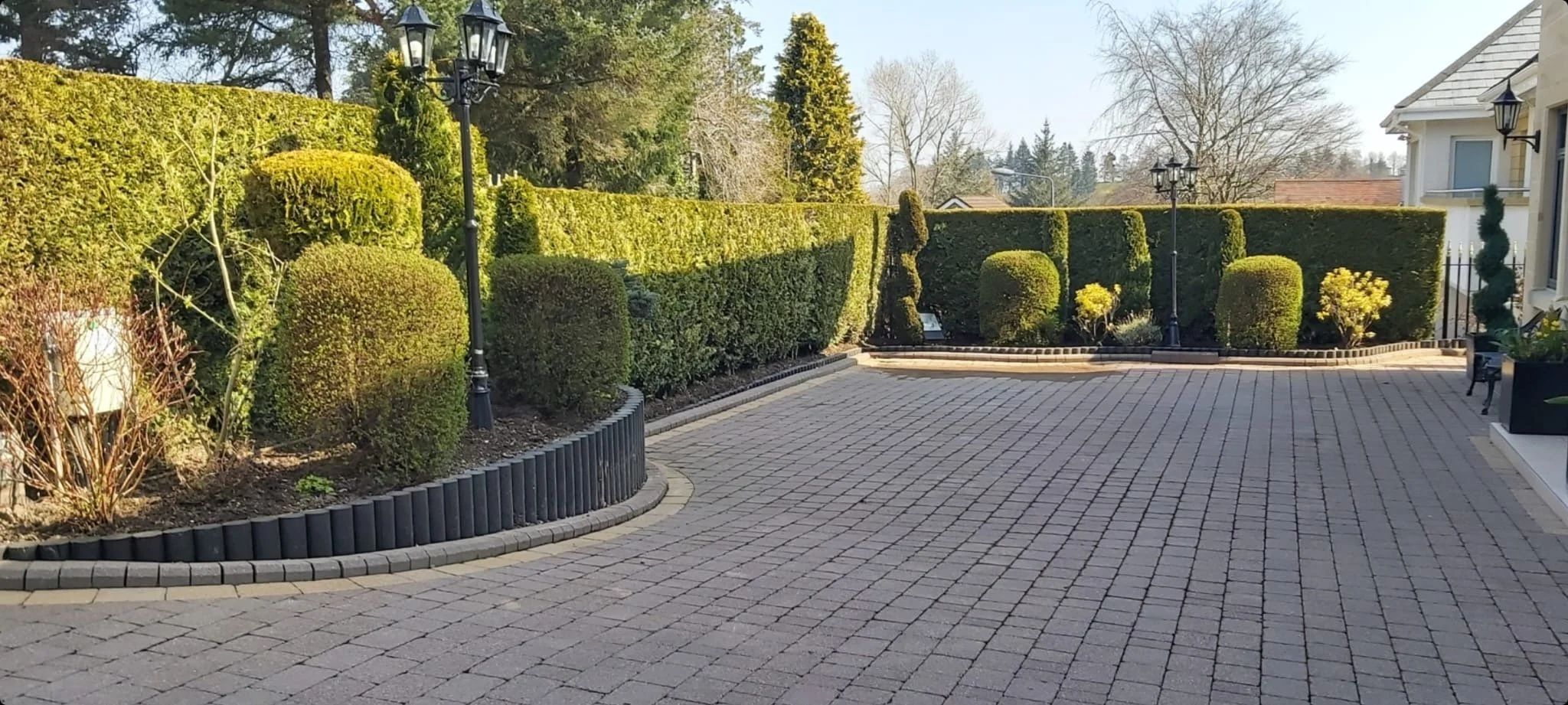 A landscaped courtyard with neatly trimmed bushes, ornamental trees, and old-fashioned black lamp posts, surrounded by a brick patio and a white house on the right side.