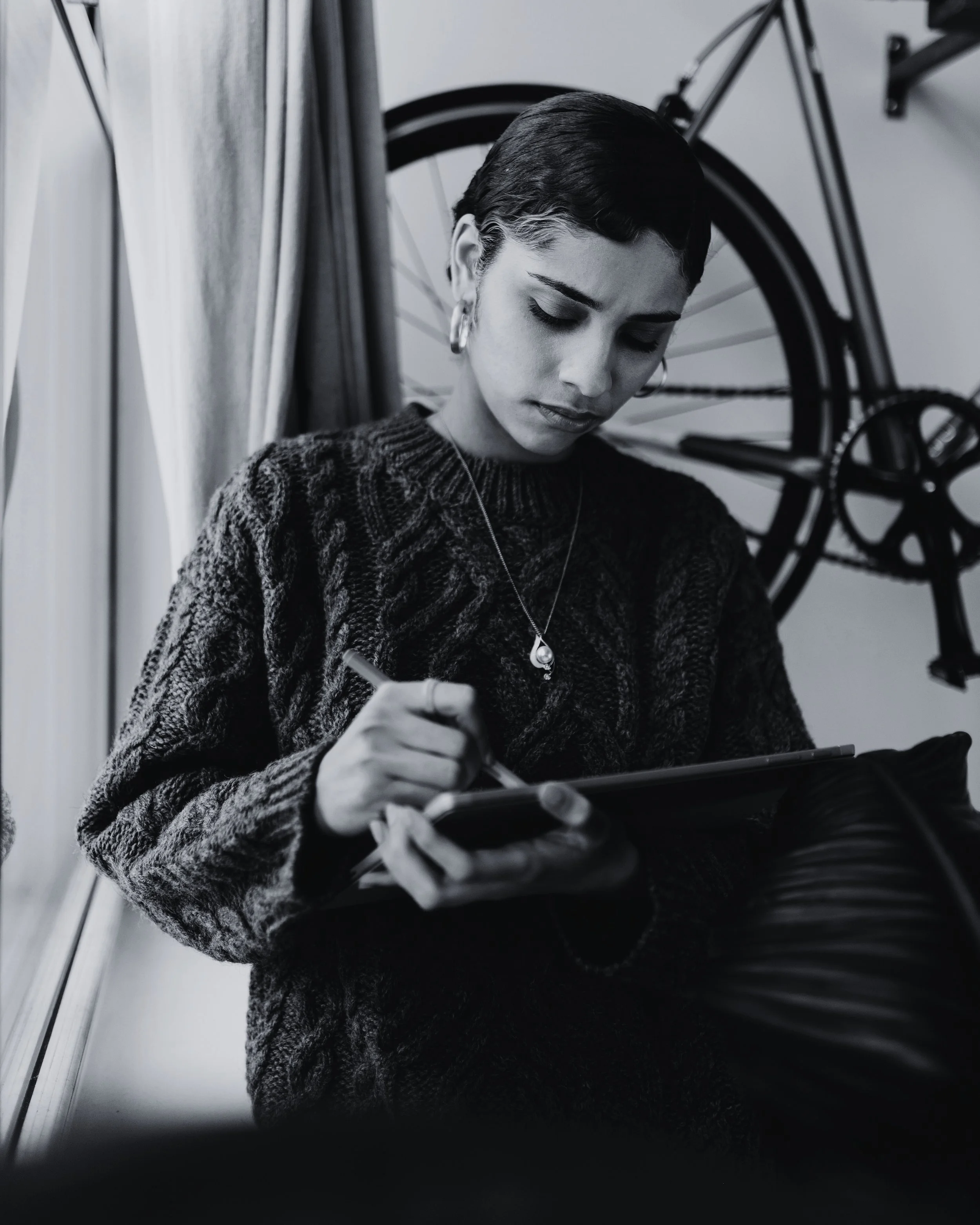 A black and white photo of a woman with short dark hair, wearing a patterned sweater and jewelry, sitting by a window with a bicycle hanging on the wall behind her, writing in a notebook.