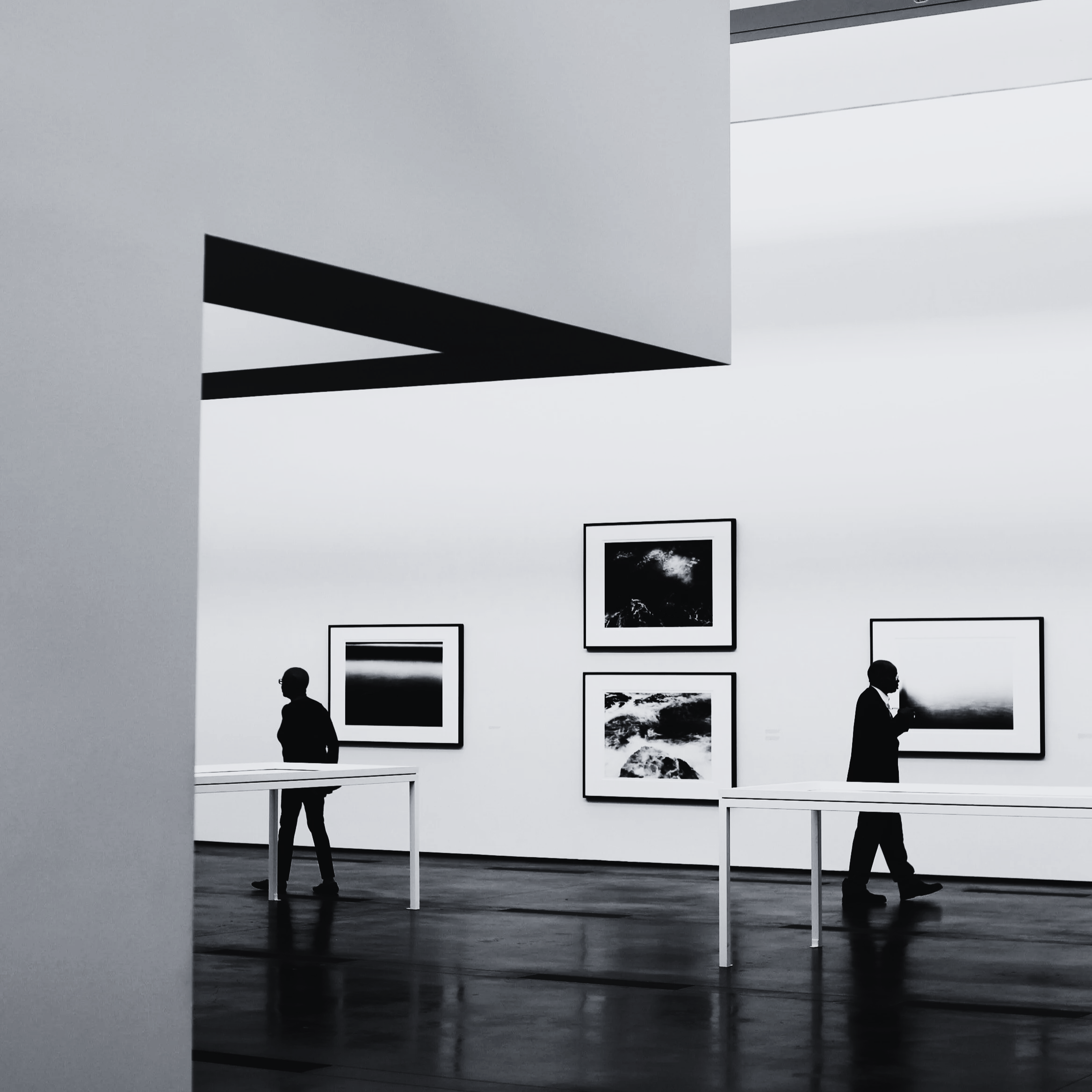 Black and white photo of an art gallery with two people viewing framed photographs on the wall. One person stands near the left wall, the other walks near the right. The gallery has white walls, dark flooring, and minimal furniture.