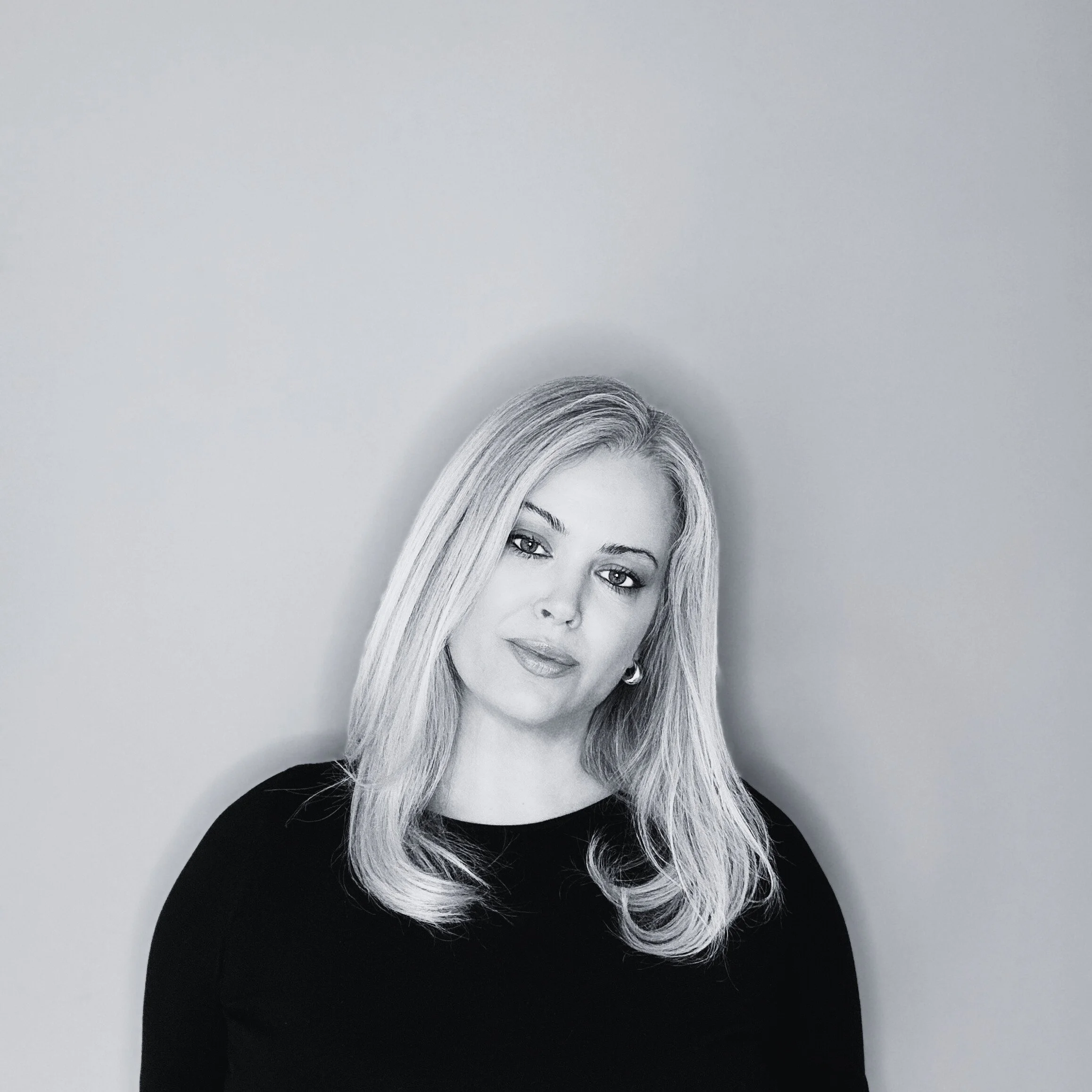 Black and white portrait of Elizabeth Elcoate, the founder of Odette, she has long, wavy blonde hair, and is wearing a black top, standing against a plain light-colored wall.