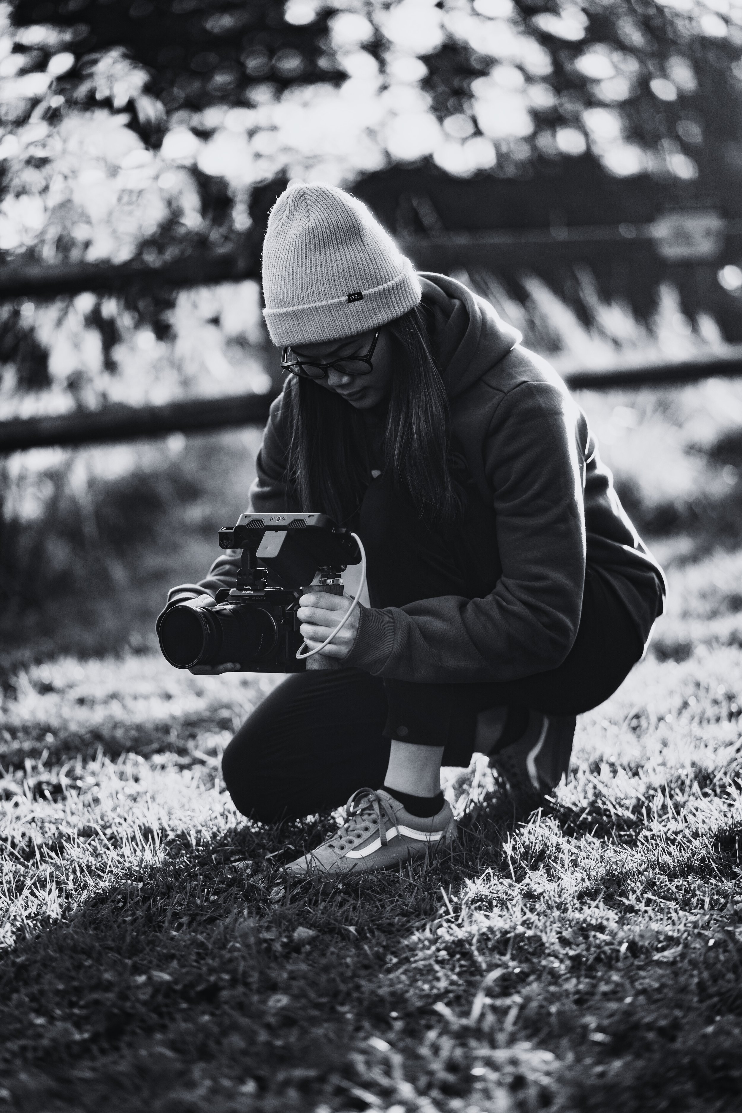 A woman wearing a beanie, glasses, hoodie, and sneakers squatting outdoors on grass while holding a camera with a stabiliser.