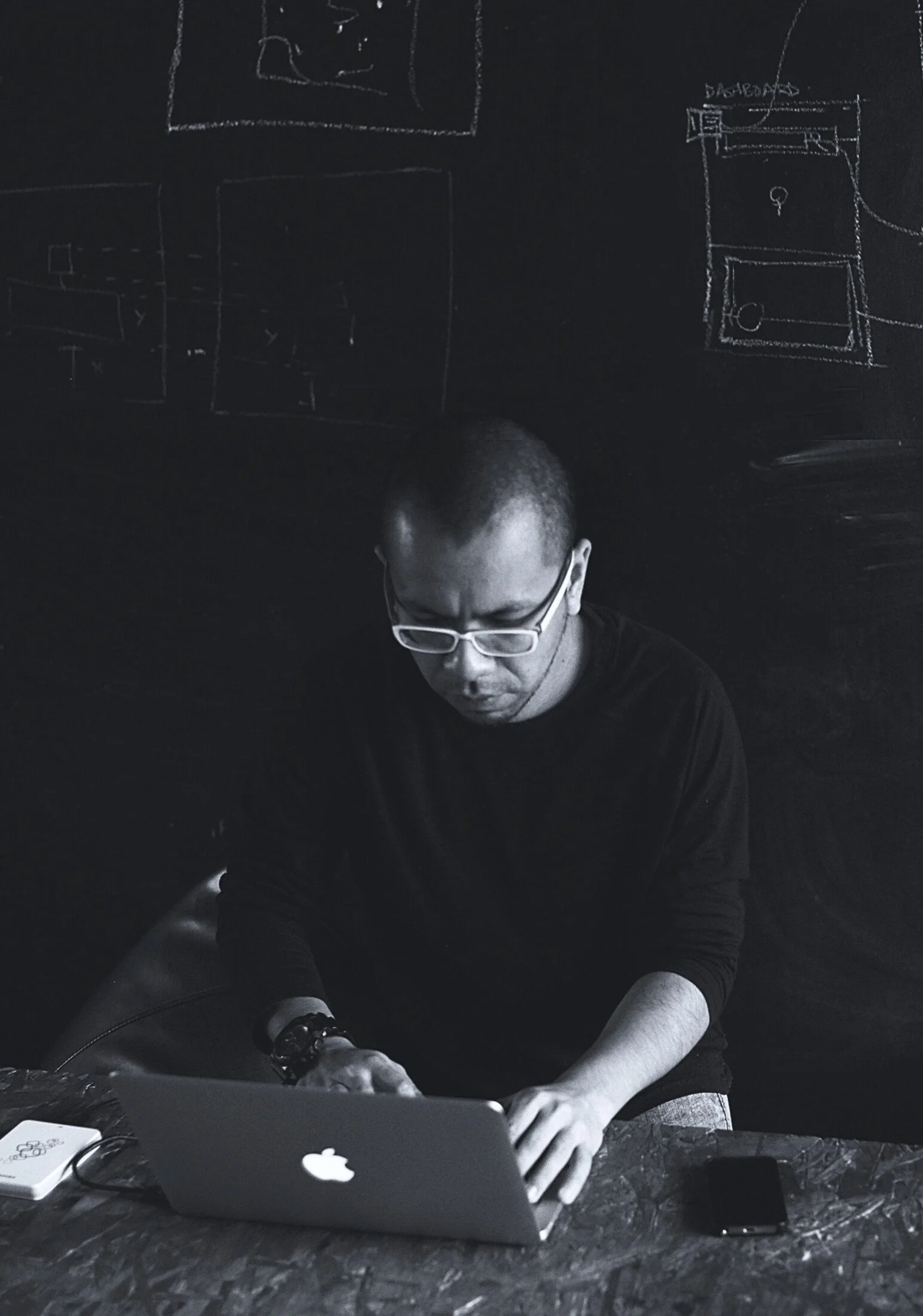 A man wearing glasses and a black shirt working on a laptop at a desk, with sketches of floor plans drawn on a blackboard behind him.
