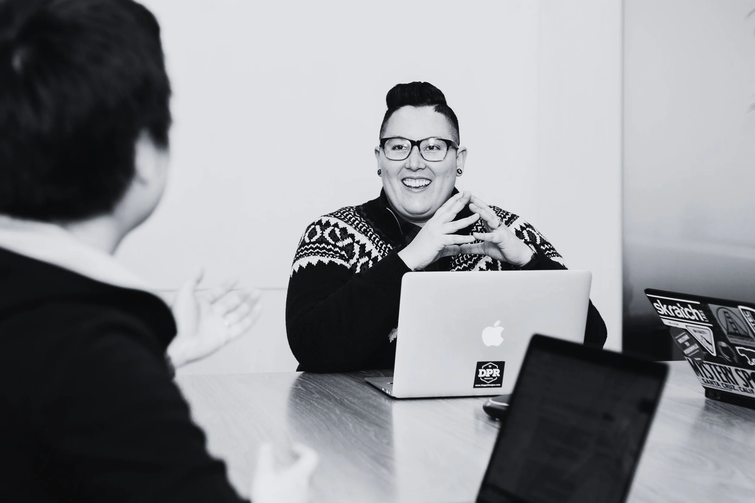 A person with glasses and short, dark hair speaking to another person in a discussion; there are laptops on the table, including a silver Apple MacBook and another with stickers, in an office setting.
