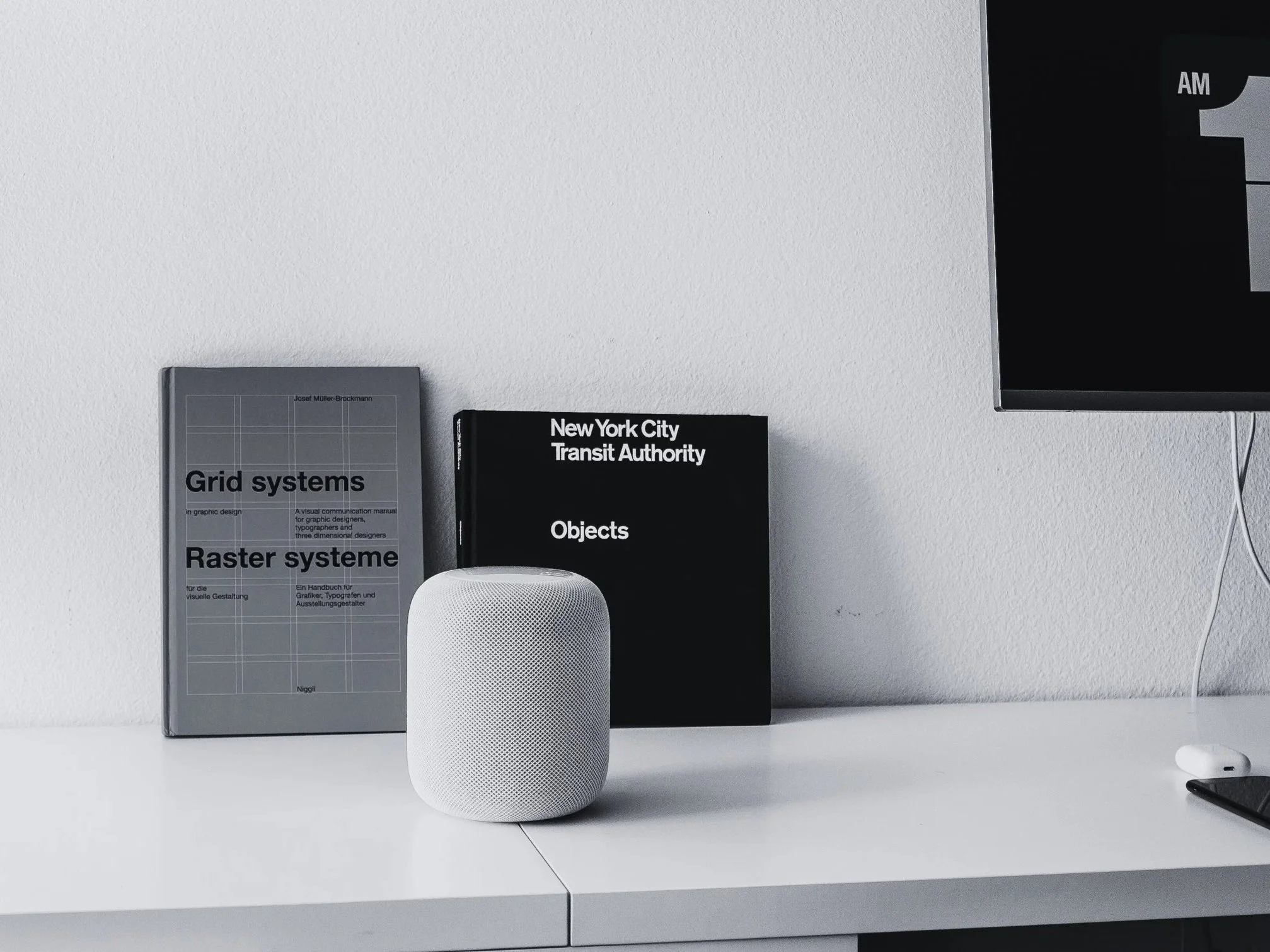 A white smart speaker on a white desk next to two books, one titled 'Grid systems' and the other 'Objects', with a black monitor displaying the time and a white remote or mouse on the desk.