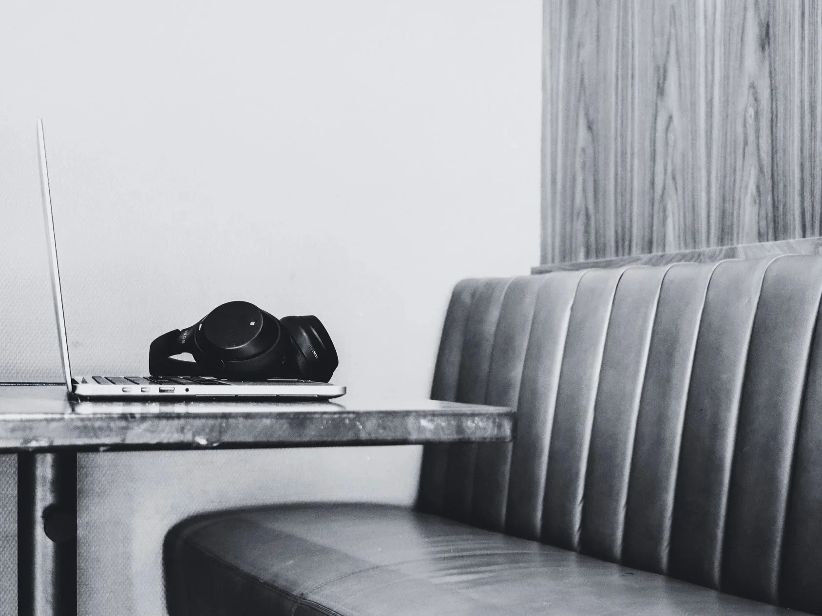 A black pair of headphones resting on a closed laptop on a table, next to a booth with a padded backrest, against a wall with wood paneling.
