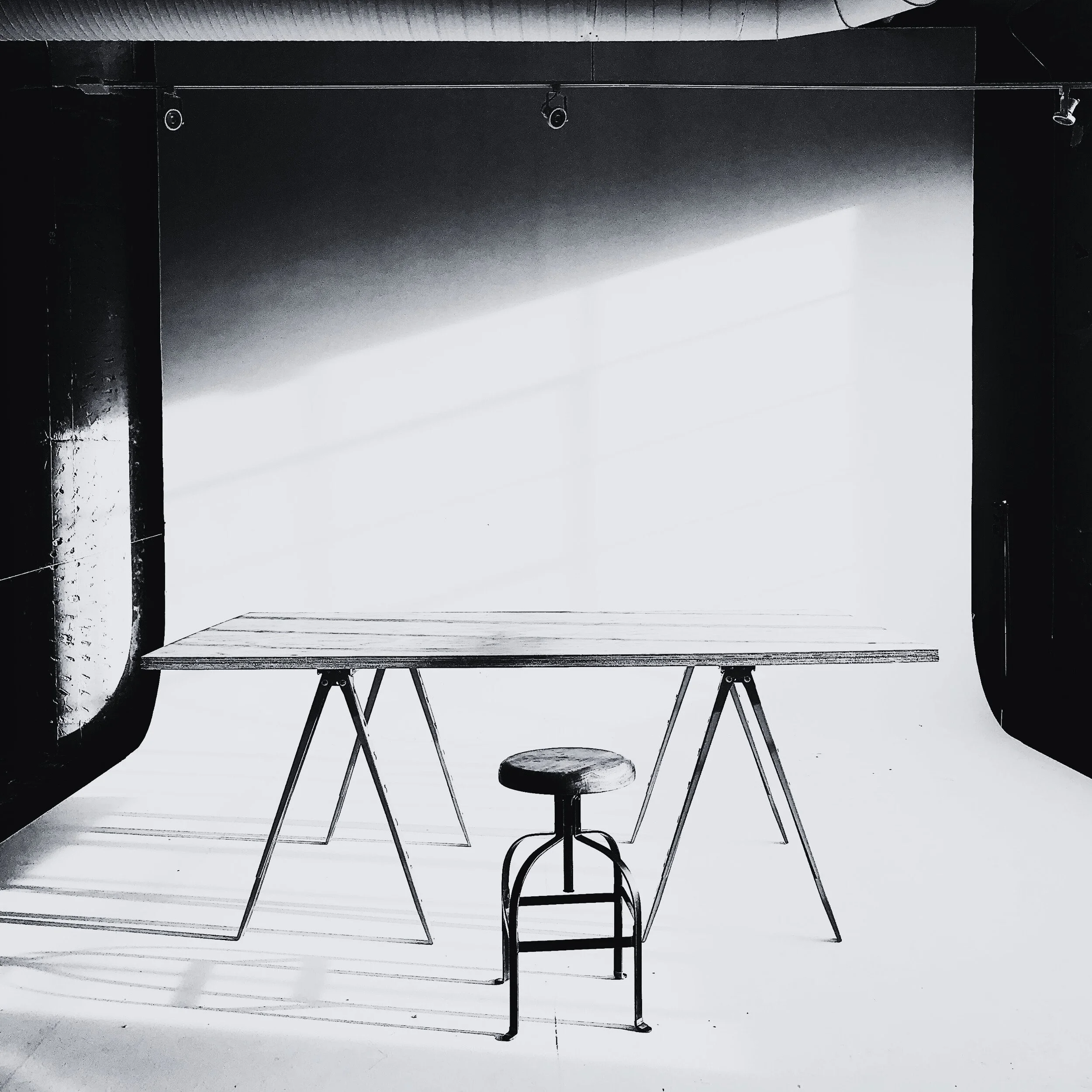A photo of a photography studio setup with a plain white backdrop, a wooden table, and a vintage wooden stool on a studio floor.