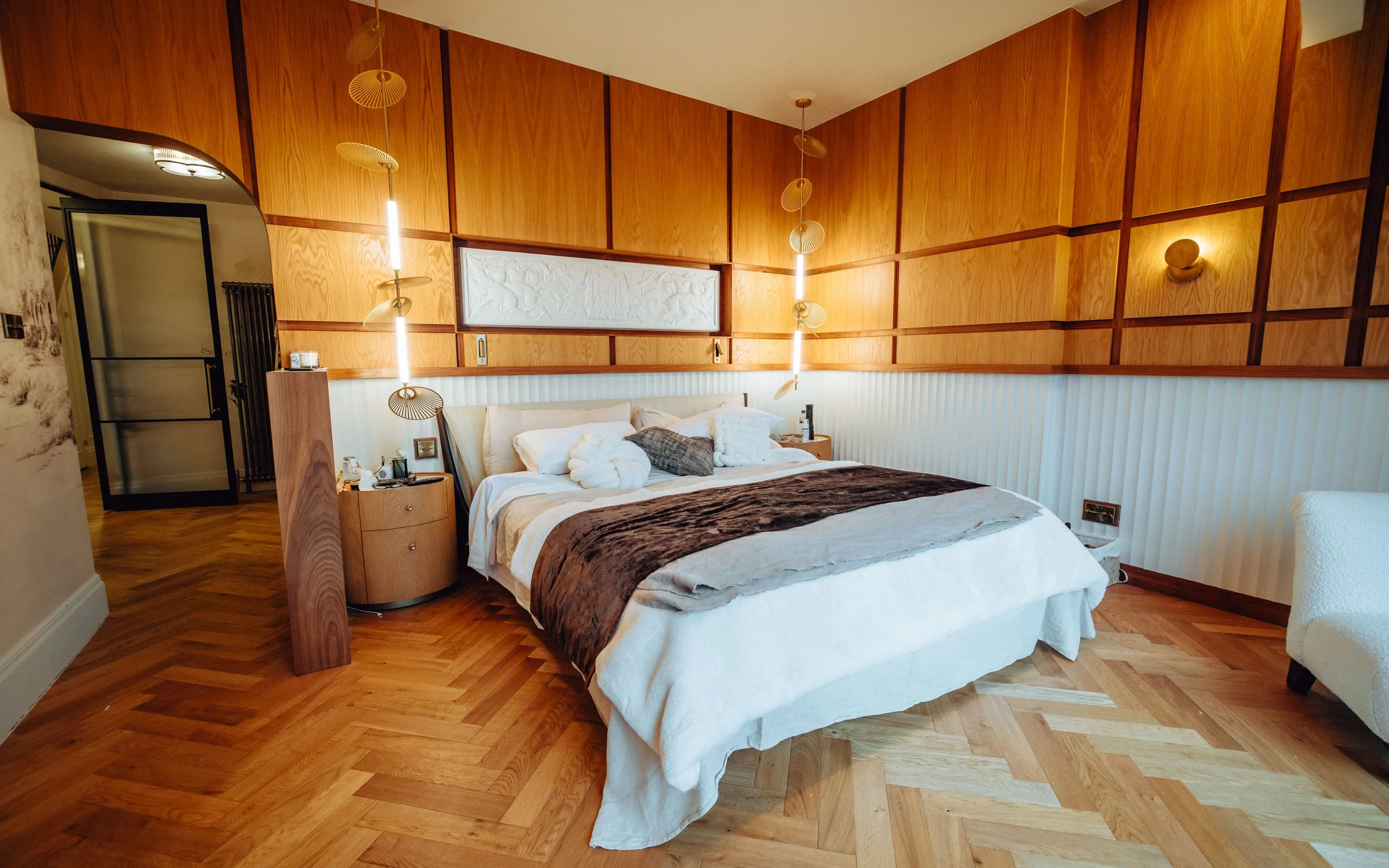 A bedroom with a wooden accent wall, white bedding, and decorative pillows, including a rolled towel shaped as a swan on the bed. There are nightstands on either side, with lamps and personal items. The room has a wood floor and modern lighting fixtures, including hanging pendant lights.