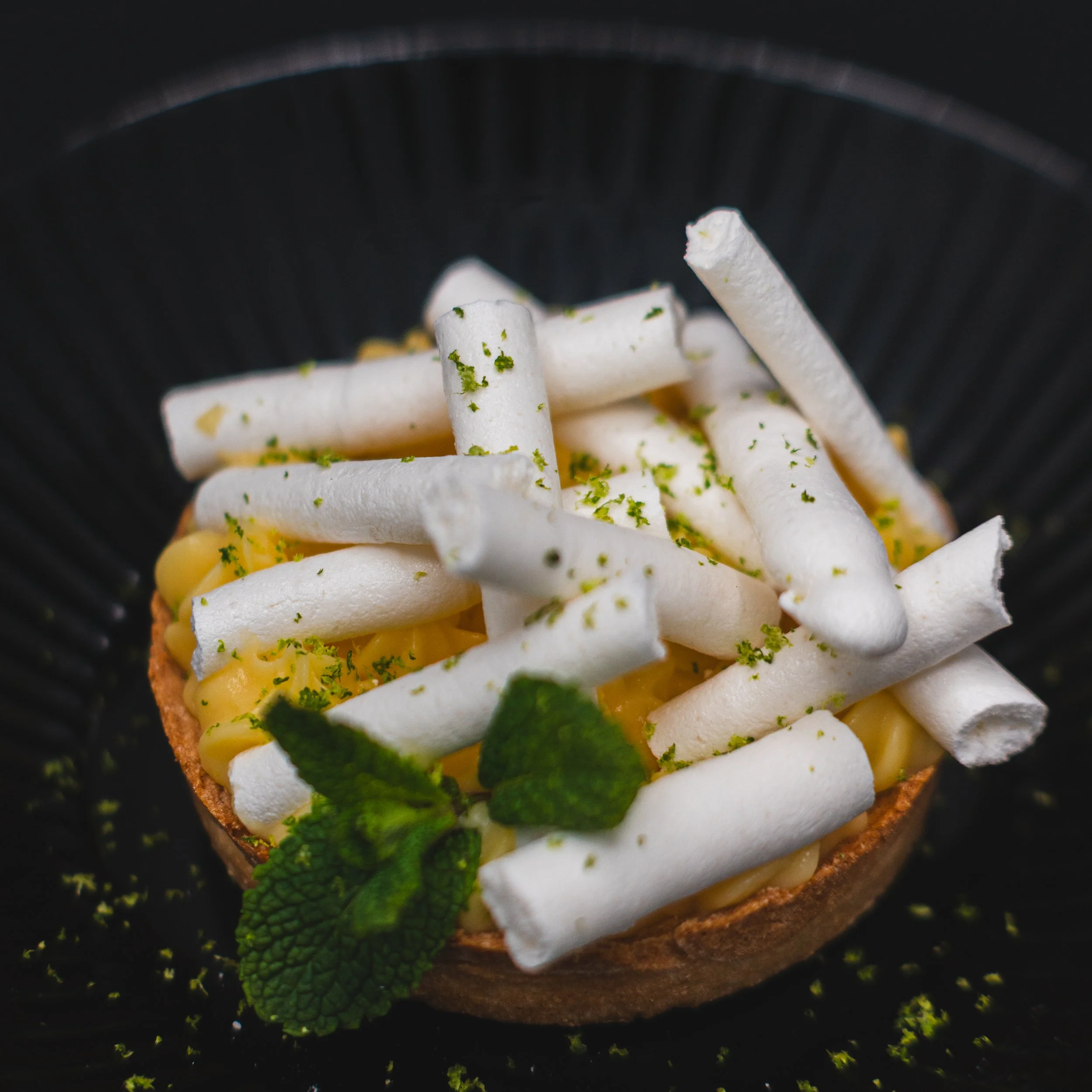 A dessert tart topped with piped white meringue sticks, lemon curd filling, garnished with fresh mint and lime zest