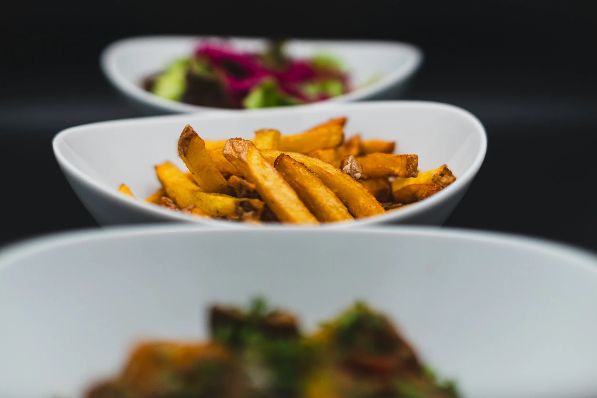 Close-up of three white bowls containing food, with French fries in the middle bowl, a salad in the bowl at the back, and an out-of-focus dish at the front.