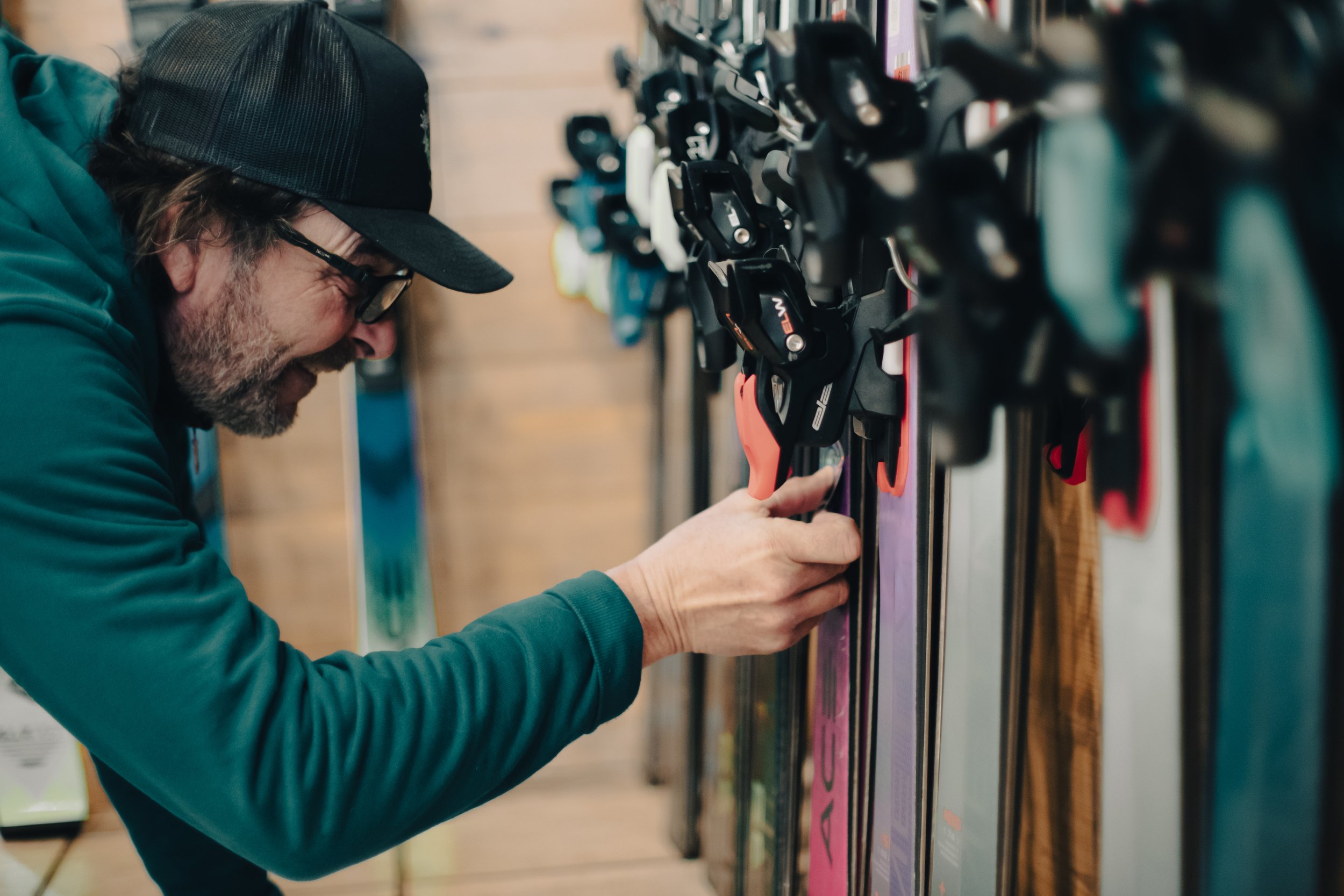 A man with glasses, wearing a cap and a green jacket, examining ski bindings on a display wall in a store.
