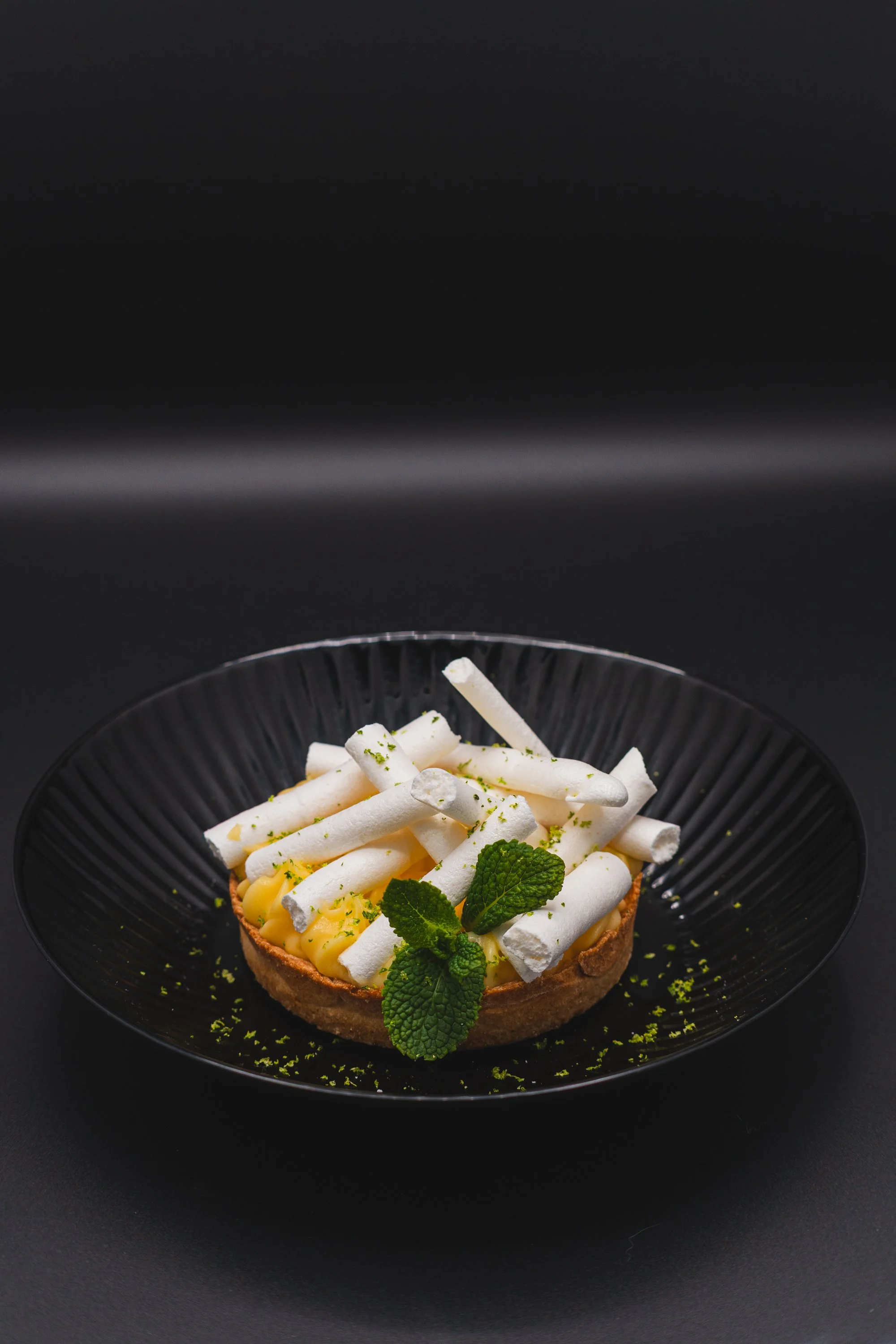 Lemon tart topped with white meringue sticks and fresh mint leaves on a black plate.