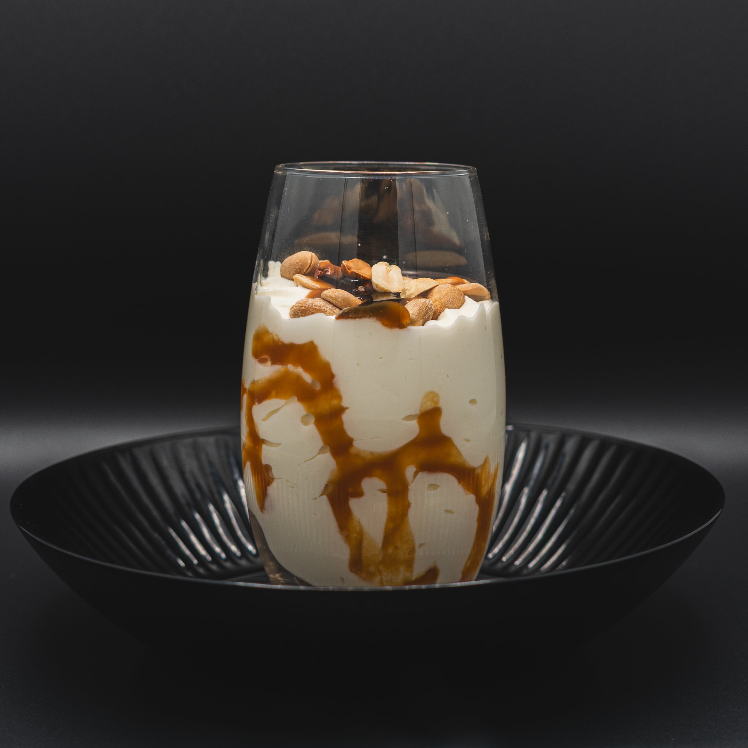 Creamy dessert in a glass with caramel drizzle, nuts, and a chocolate topping, served on a black plate against a dark background.