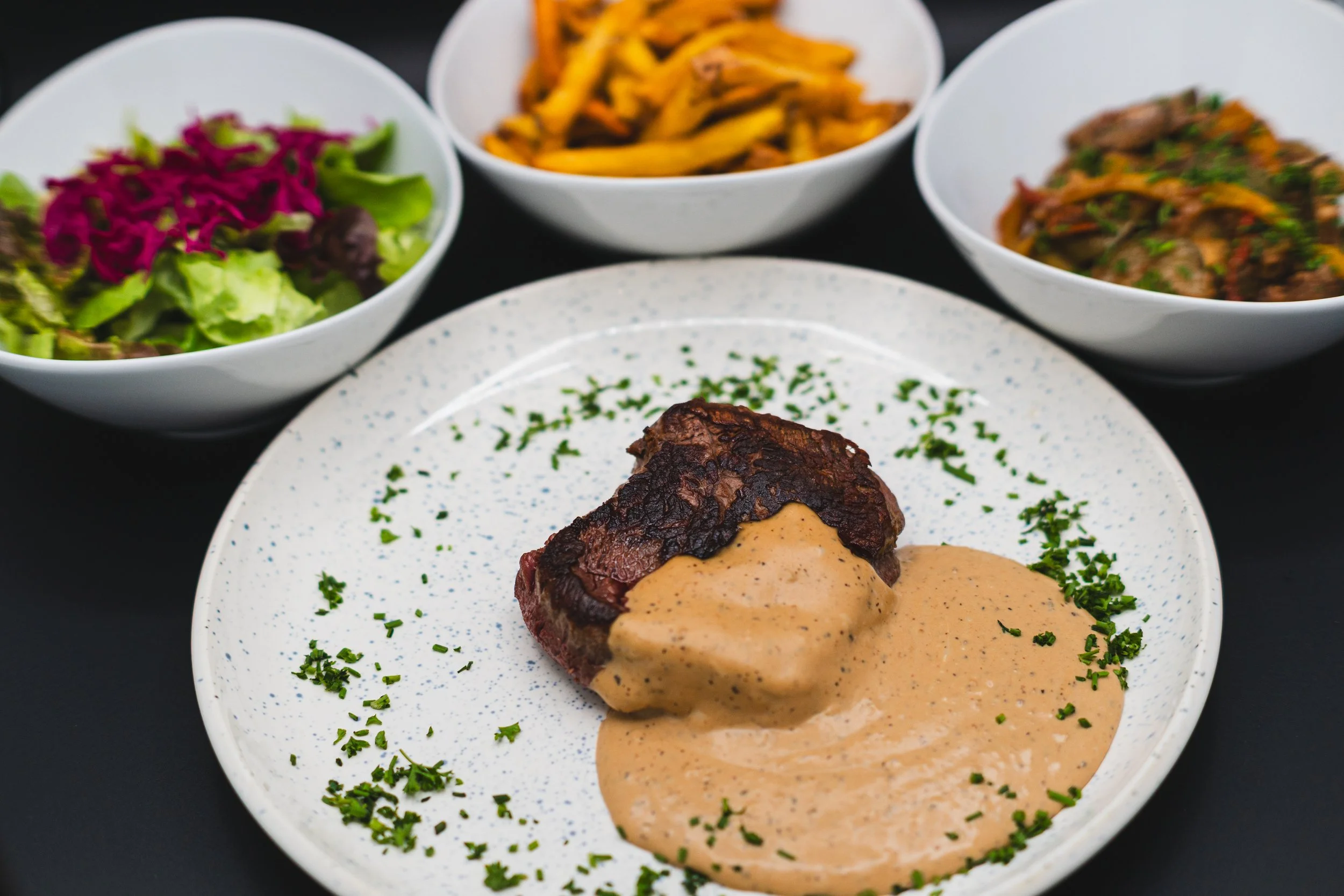 A plate with a cooked steak topped with creamy sauce, garnished with chopped herbs, and three bowls of side dishes including a salad, sweet potato fries, and a vegetable medley.