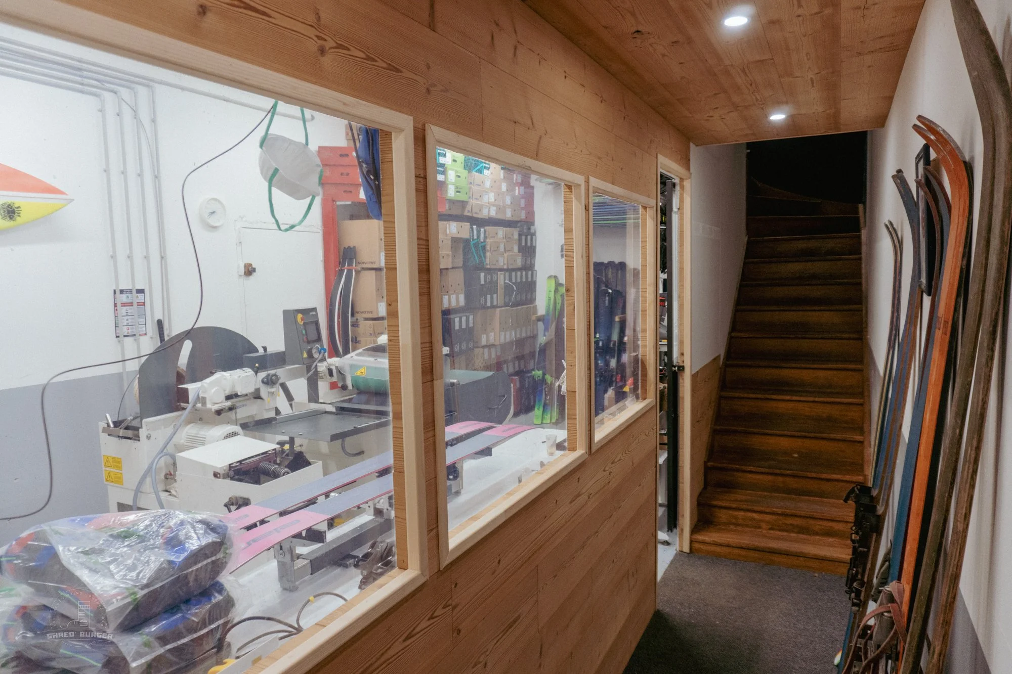 View of a ski and snowboard shop with a staircase, skis, and snowboards on the wall and a window looking into a storage room with ski equipment.