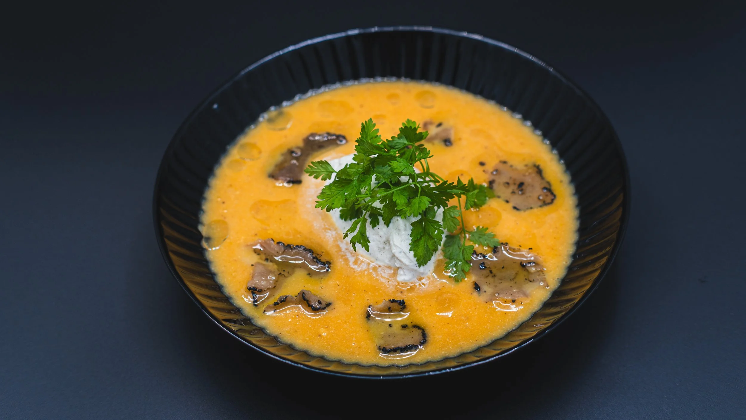 A black bowl containing a creamy yellow soup garnished with a sprig of fresh parsley and dollops of sour cream, with pieces of black truffle on top, placed on a dark surface.
