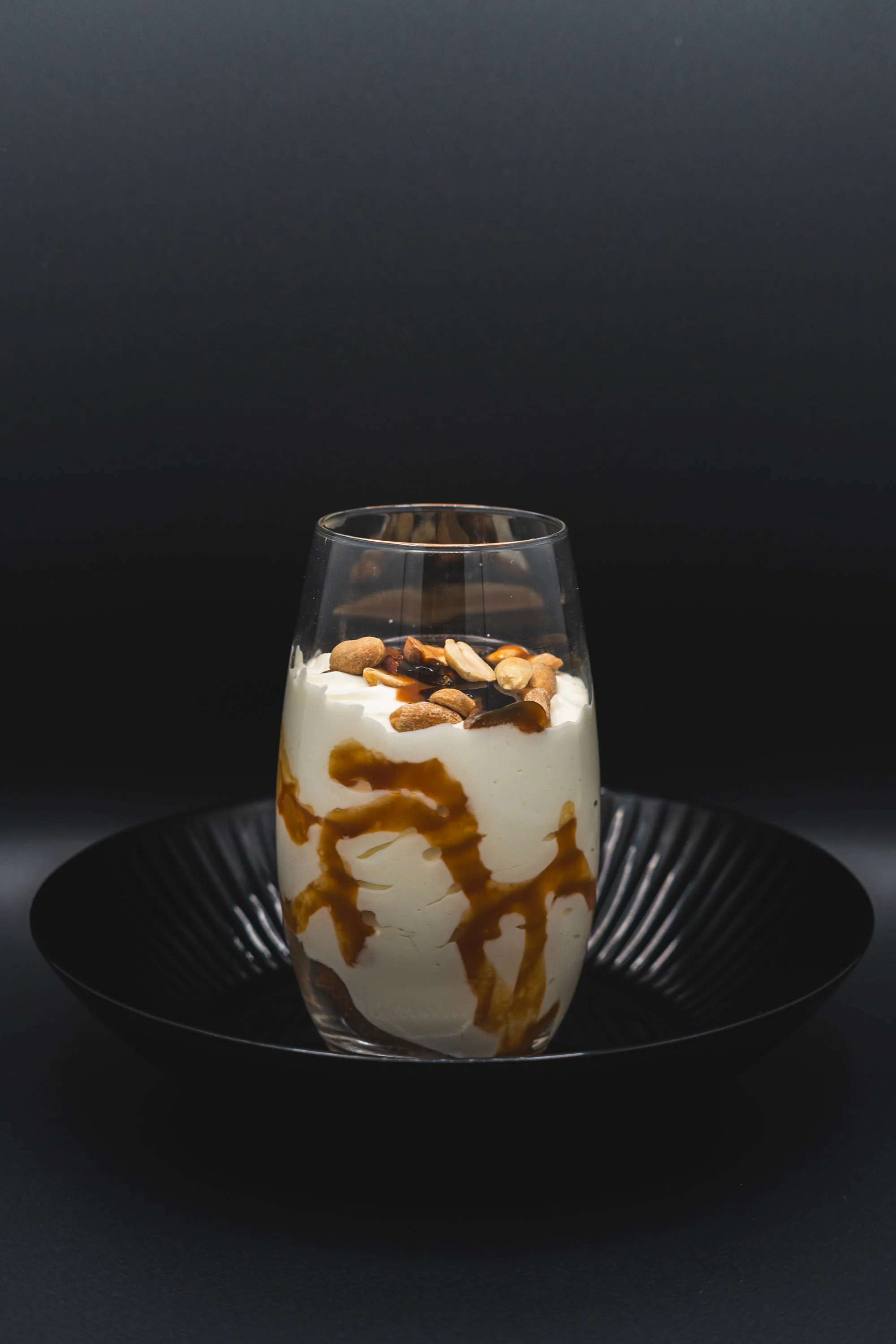 Dessert in a clear glass garnished with nuts and drizzled with caramel sauce, placed on a black plate against a dark background.