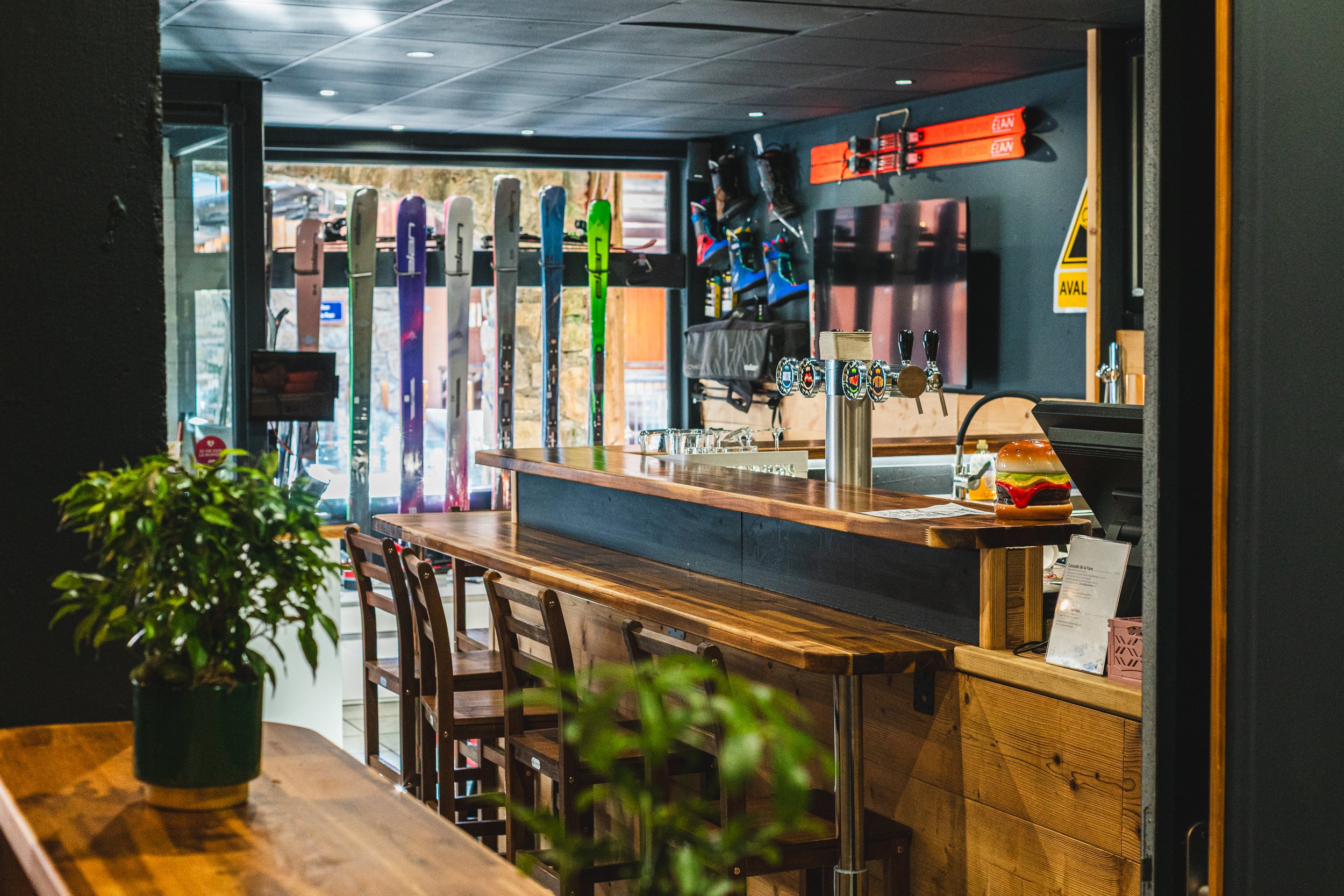 Interior view of a ski lodge bar area with a wooden countertop, colorful skis on display, a row of beer taps, and a burger on the counter.
