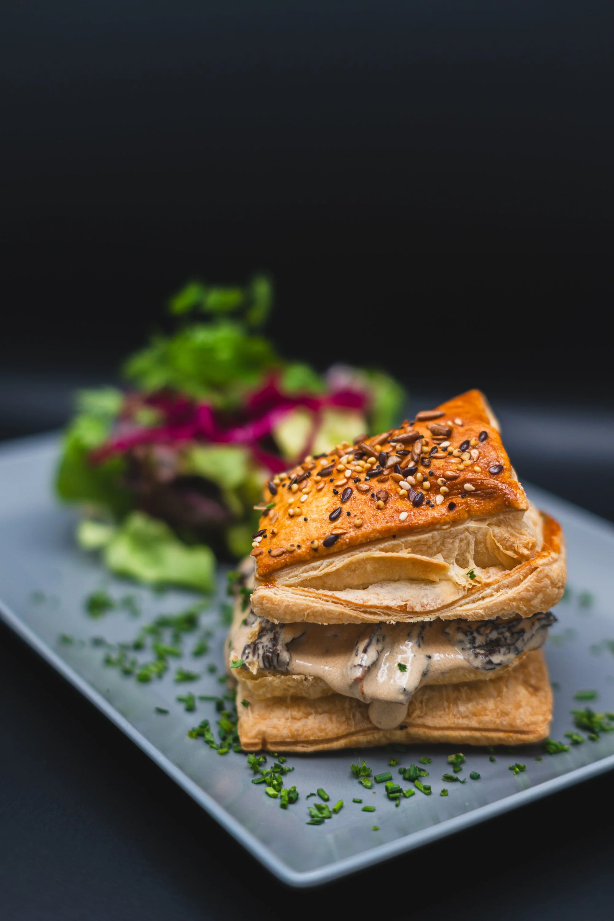 A savory pastry sandwich with a flaky, golden-brown crust topped with sesame seeds and black seeds, filled with creamy mushrooms, served on a white plate with a side salad of mixed greens and purple lettuce.