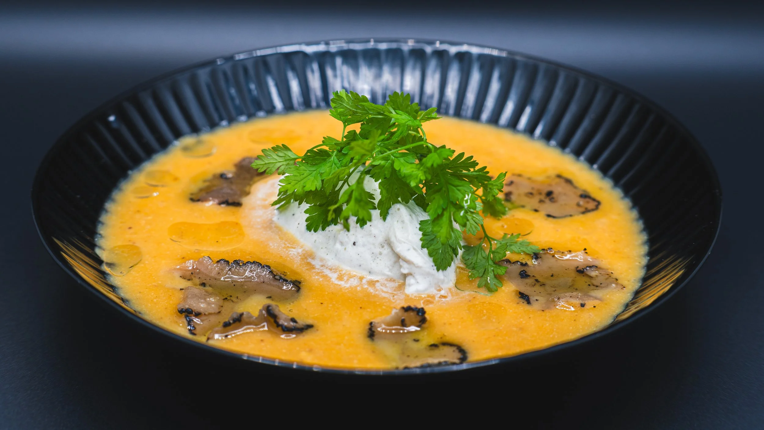 A black bowl of creamy orange soup garnished with chopped herbs and a dollop of sour cream, topped with fresh parsley and sliced mushrooms.