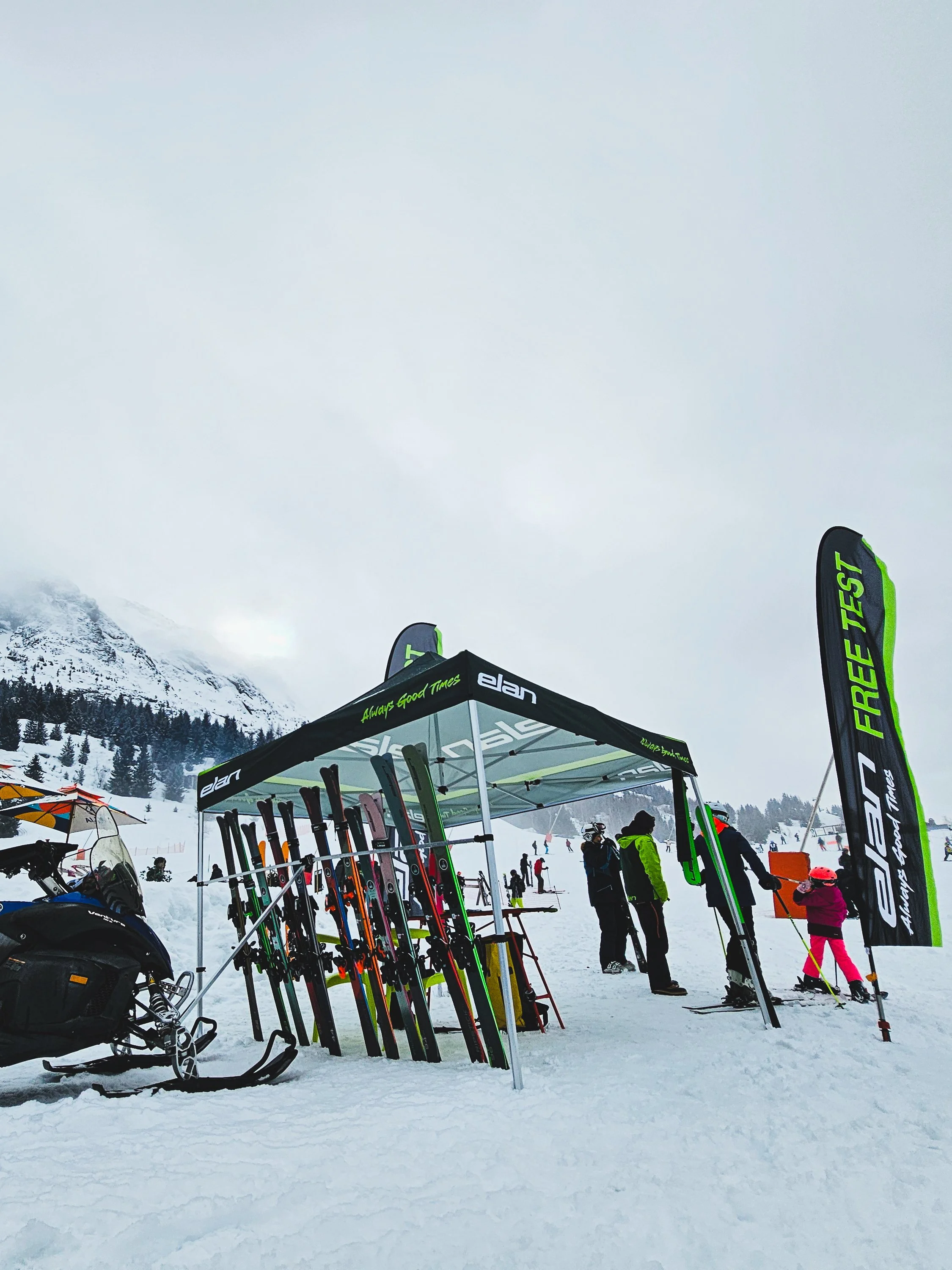 Ski rental booth on a snowy mountain with skis and snowboards, and people in winter gear.