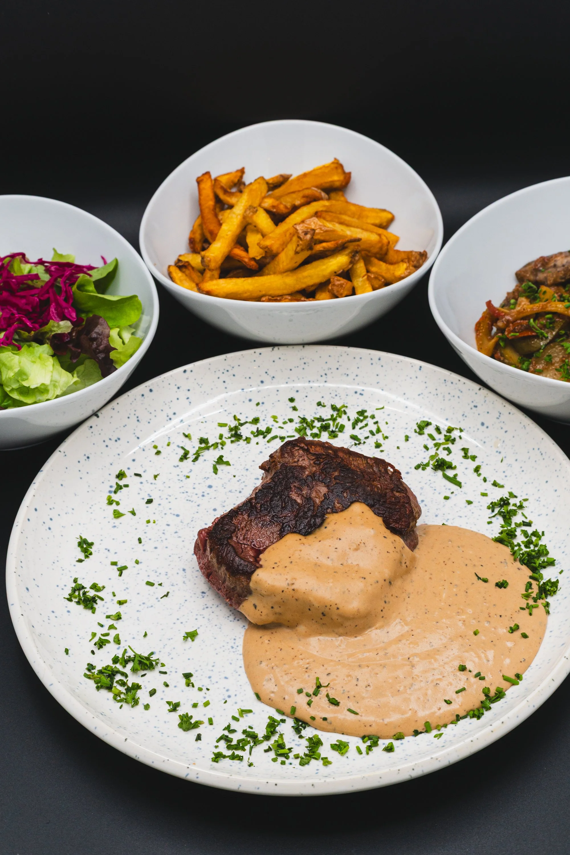 A plate with a cooked steak topped with creamy sauce and garnished with chopped parsley, surrounded by four bowls of side dishes including salad, French fries, vegetables, and cooked meat.