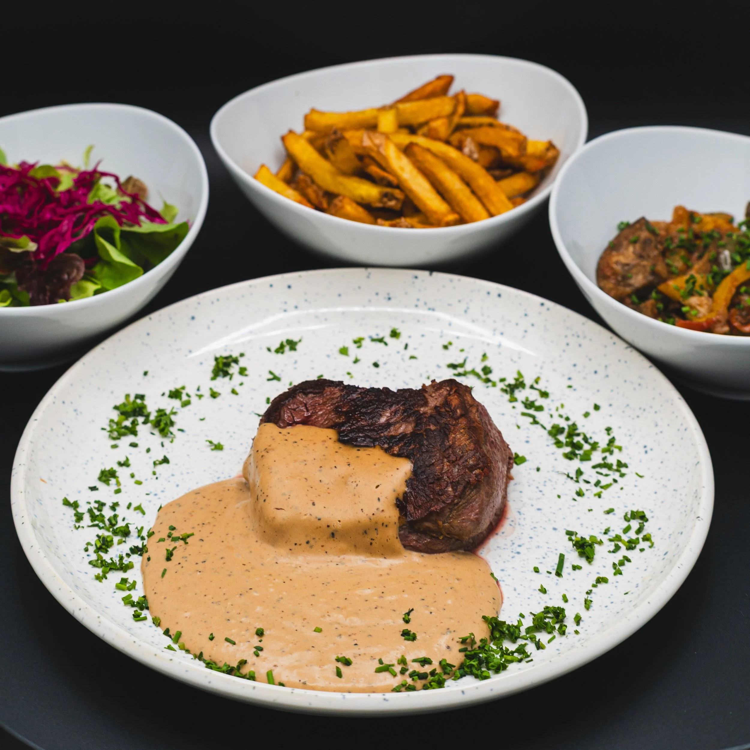 A plated steak topped with creamy sauce, garnished with chopped parsley, surrounded by side dishes of fries, salad, and roasted vegetables in white bowls.