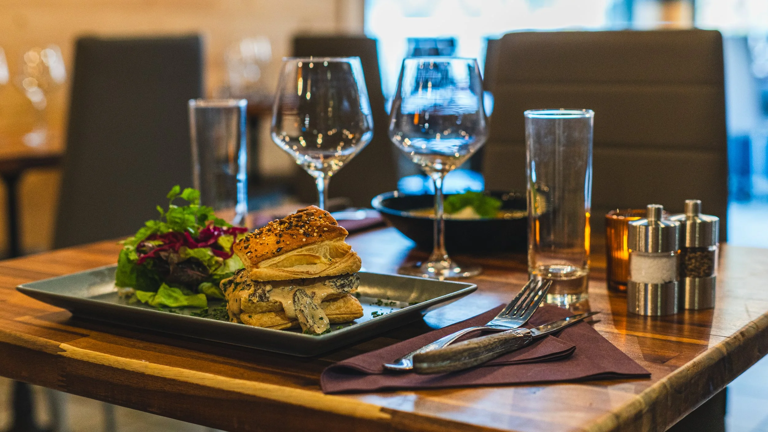 A plate with a sandwich and salad on a restaurant table, set with wine glasses, water glasses, cutlery, and salt and pepper shakers.
