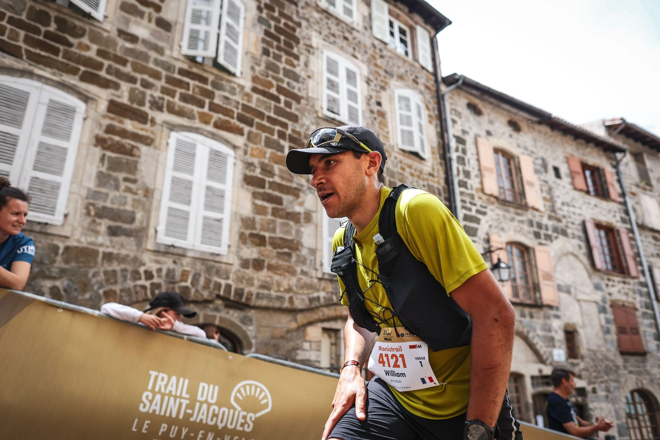 Un homme coureur portant un maillot jaune, un gilet de course et une casquette, semble épuisé ou en effort intense lors d'une course dans une rue pavée, avec un bâtiment en pierre en arrière-plan, et d'autres spectateurs ou participants visibles.