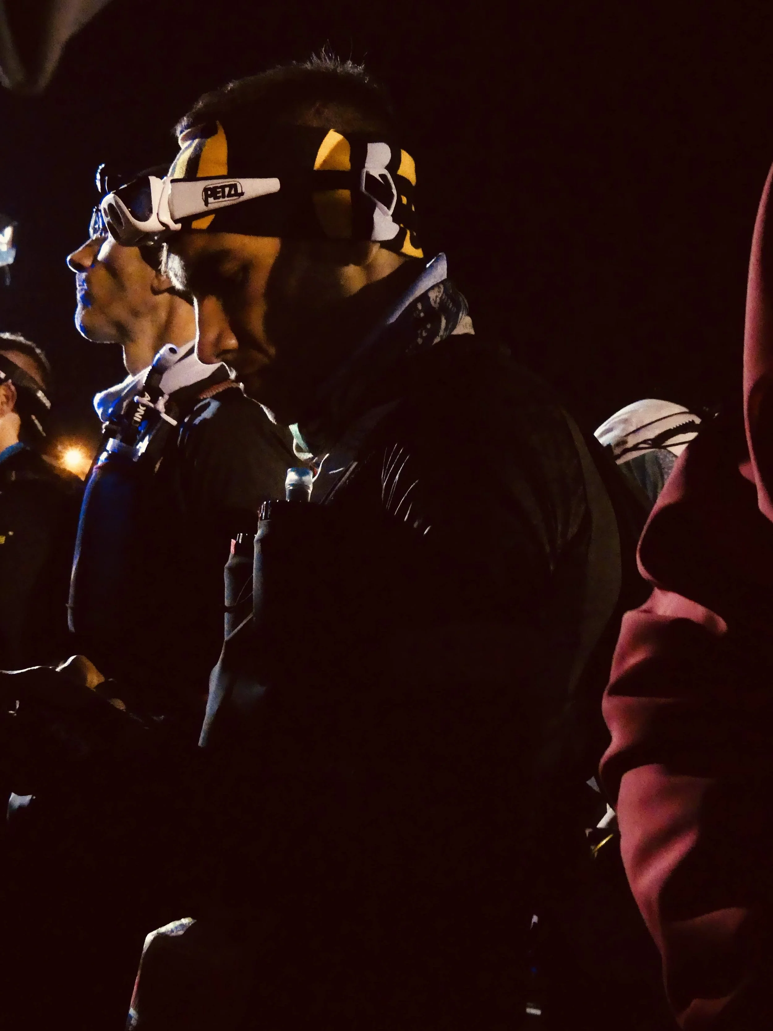 Groupe de personnes, probablement des grimpeurs ou des alpinistes, portant des équipements pour la randonnée ou l'escalade, avec toile de fond nocturne.