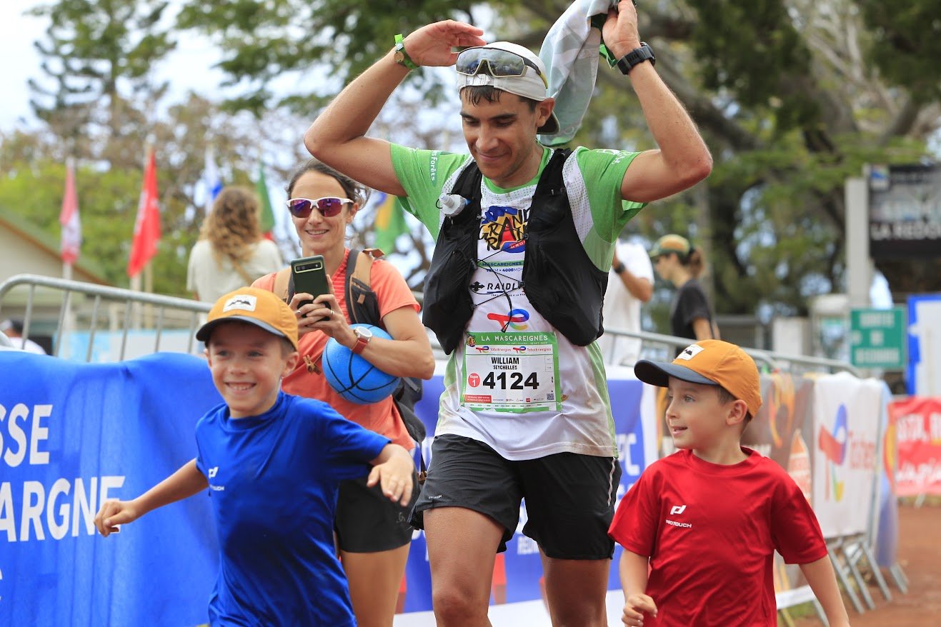Un groupe de personnes participant à une course ou un marathon, avec un homme en montre, deux jeunes garçons et deux femmes, dans un environnement extérieur avec des arbres et des barrières de sécurité.