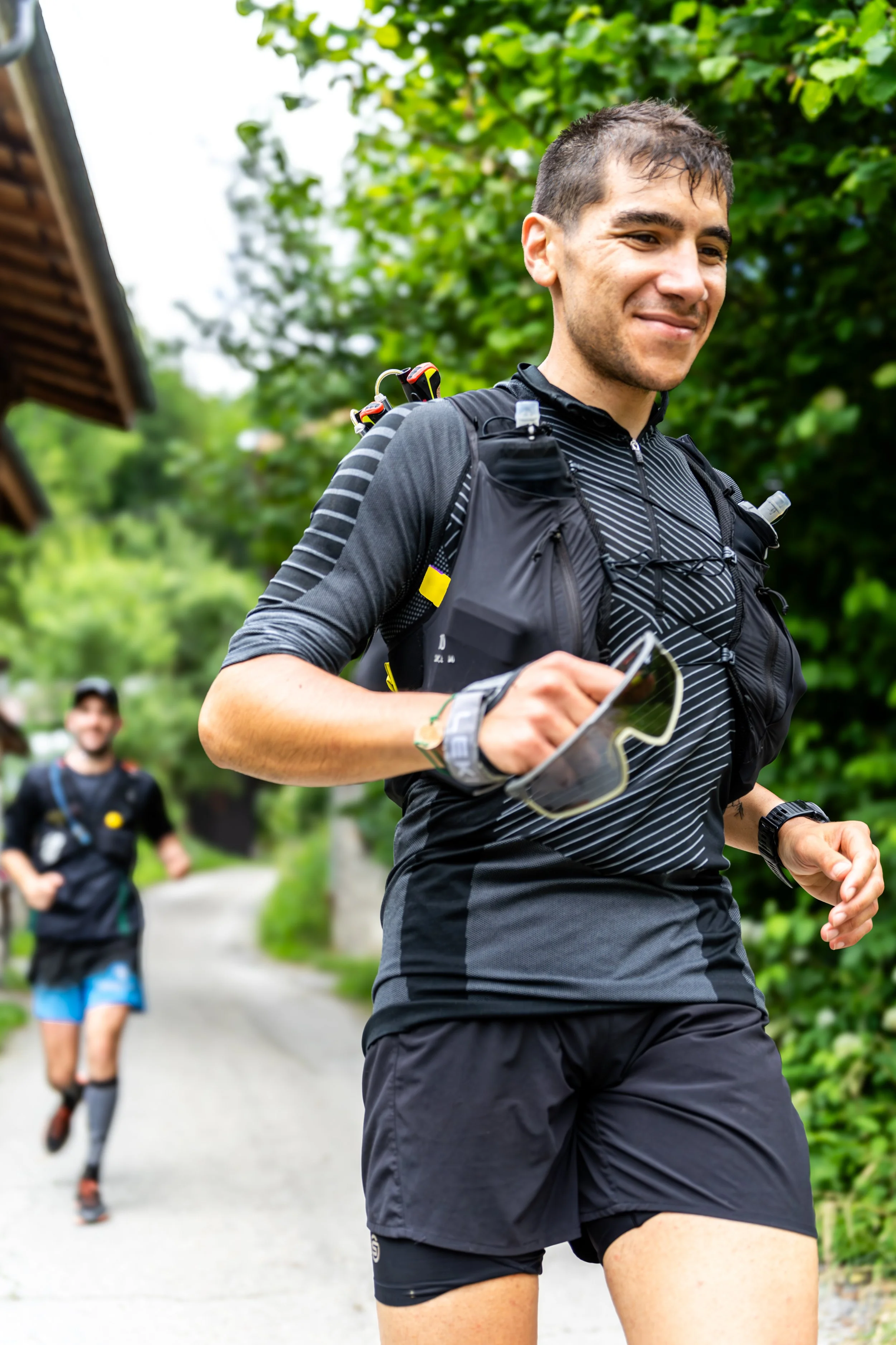 Jeune homme faisant de la course en plein air, portant une tenue de course, un sac à dos, et tenant des lunettes de soleil, avec un autre coureur à l'arrière, dans un environnement vert et boisé.