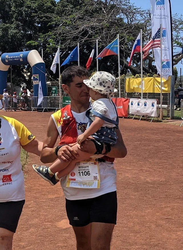 Un homme tenant un jeune enfant dans un terrain de course, avec des drapeaux nationaux en arrière-plan.