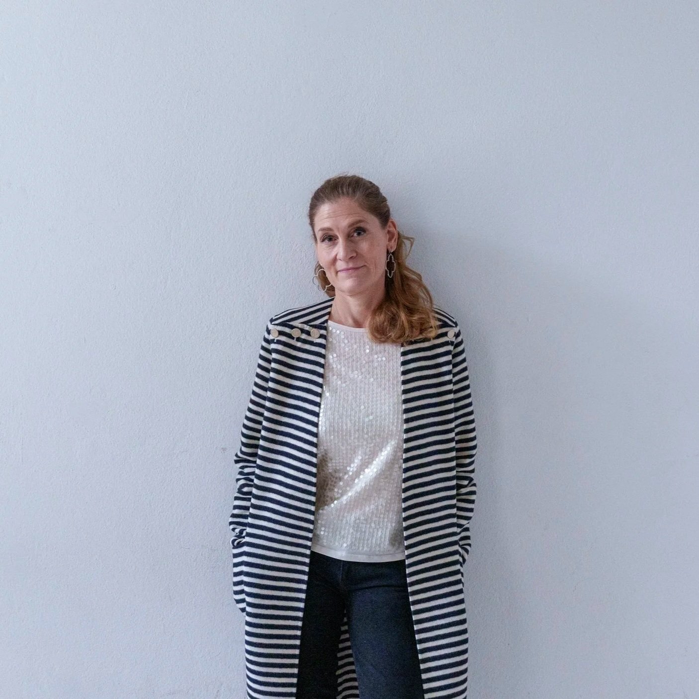 A woman wearing a striped blazer and a white sequin top standing against a plain light-colored wall.