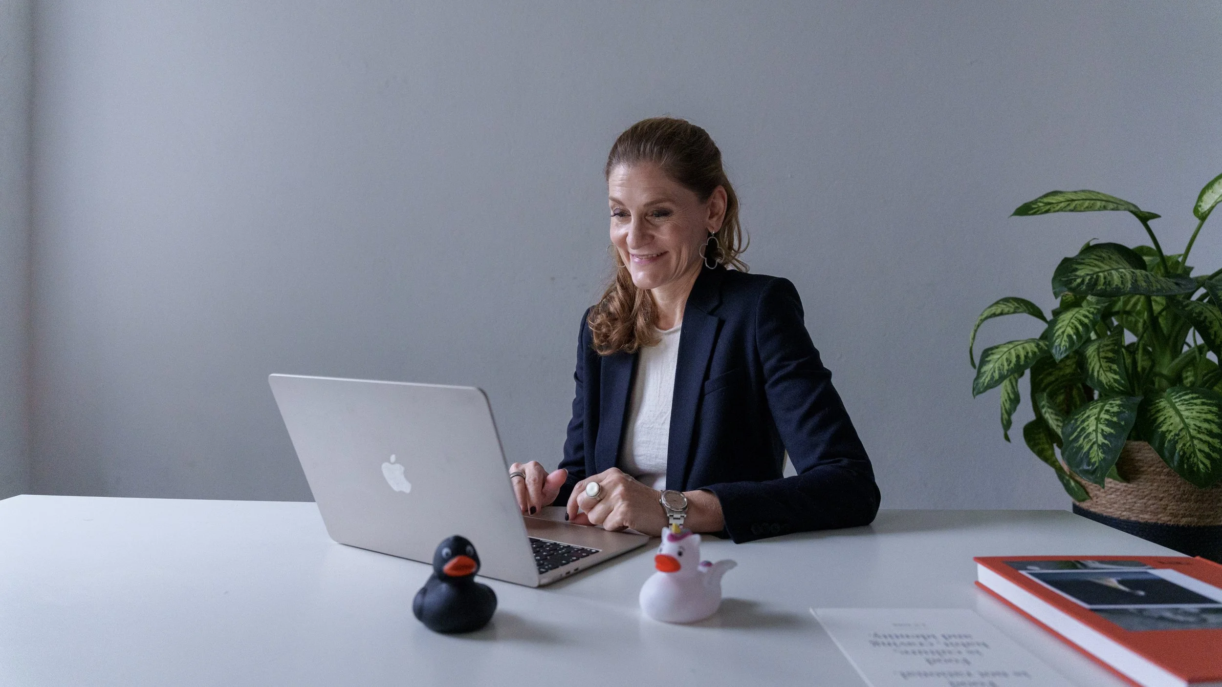 A woman in a navy blazer and white shirt sits at a white desk, working on a silver MacBook. She has long, wavy hair and is smiling while looking at the laptop. On the desk are two rubber duck figures, a black one and a white one, and a red book. A large green potted plant is to her right against a gray wall.