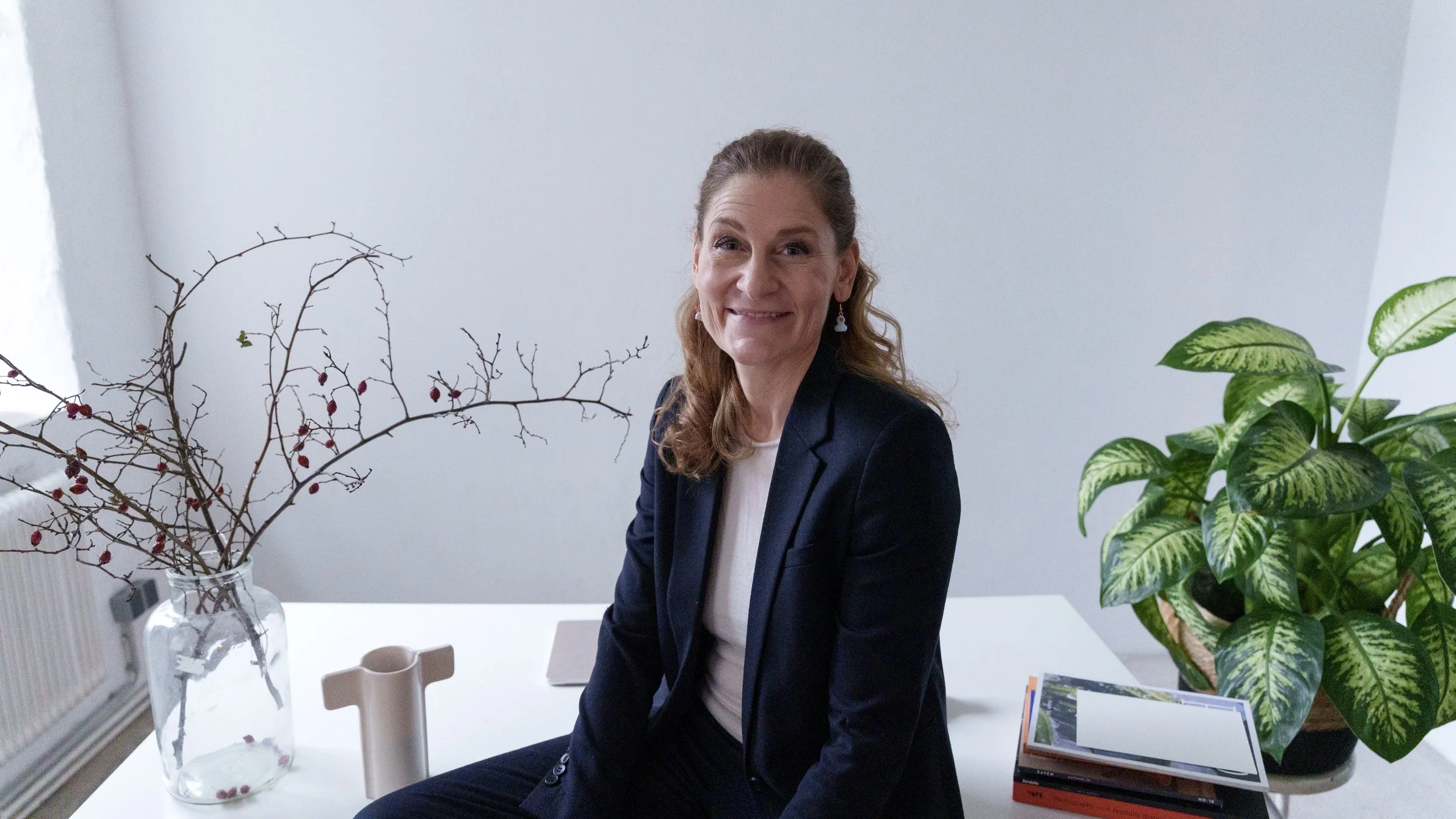 A woman with long, wavy hair wearing a navy blazer and white top, sitting at a white table with a glass vase containing with bare twigs with red berries, a large green plant, and a stack of books.